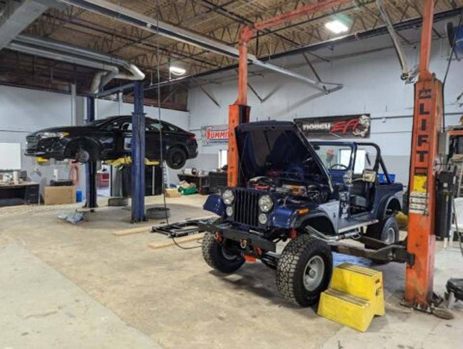 A blue Jeep with its hood open and a black car on lifts inside a garage.