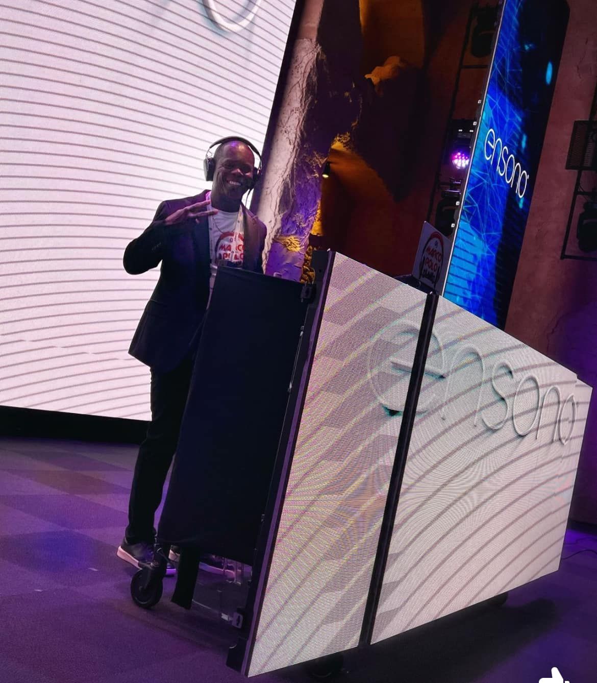DJ wearing headphones and a suit, smiling and gesturing, standing in front of a DJ booth with LED panels displaying white wave patterns.