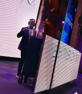 DJ in a dark suit with headphones on, standing next to a light-up DJ booth, smiling and gesturing with one hand. Interior setting.