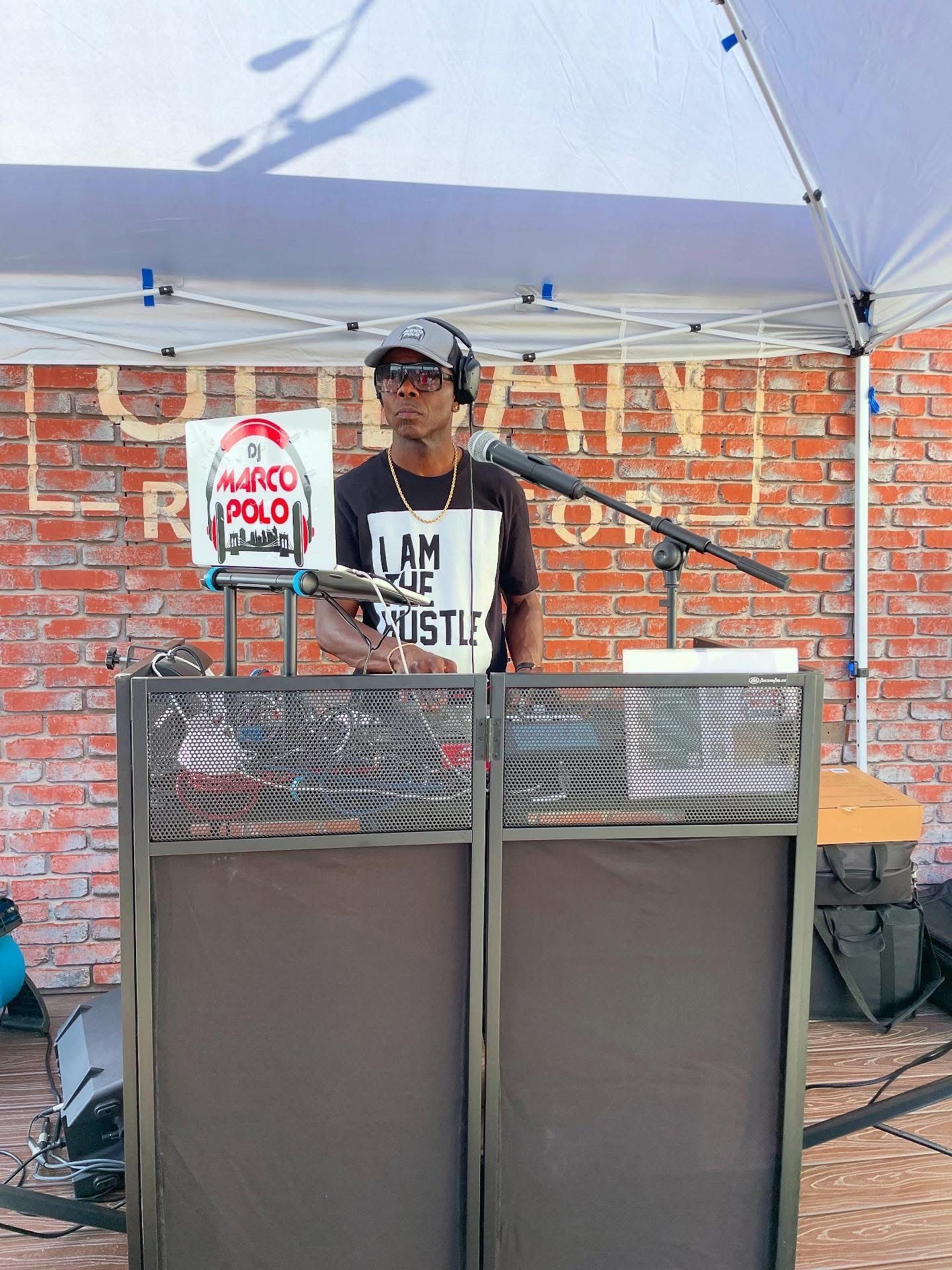 DJ at an outdoor event, standing behind equipment, wearing headphones and a t-shirt. A tent provides shade against a brick wall background.
