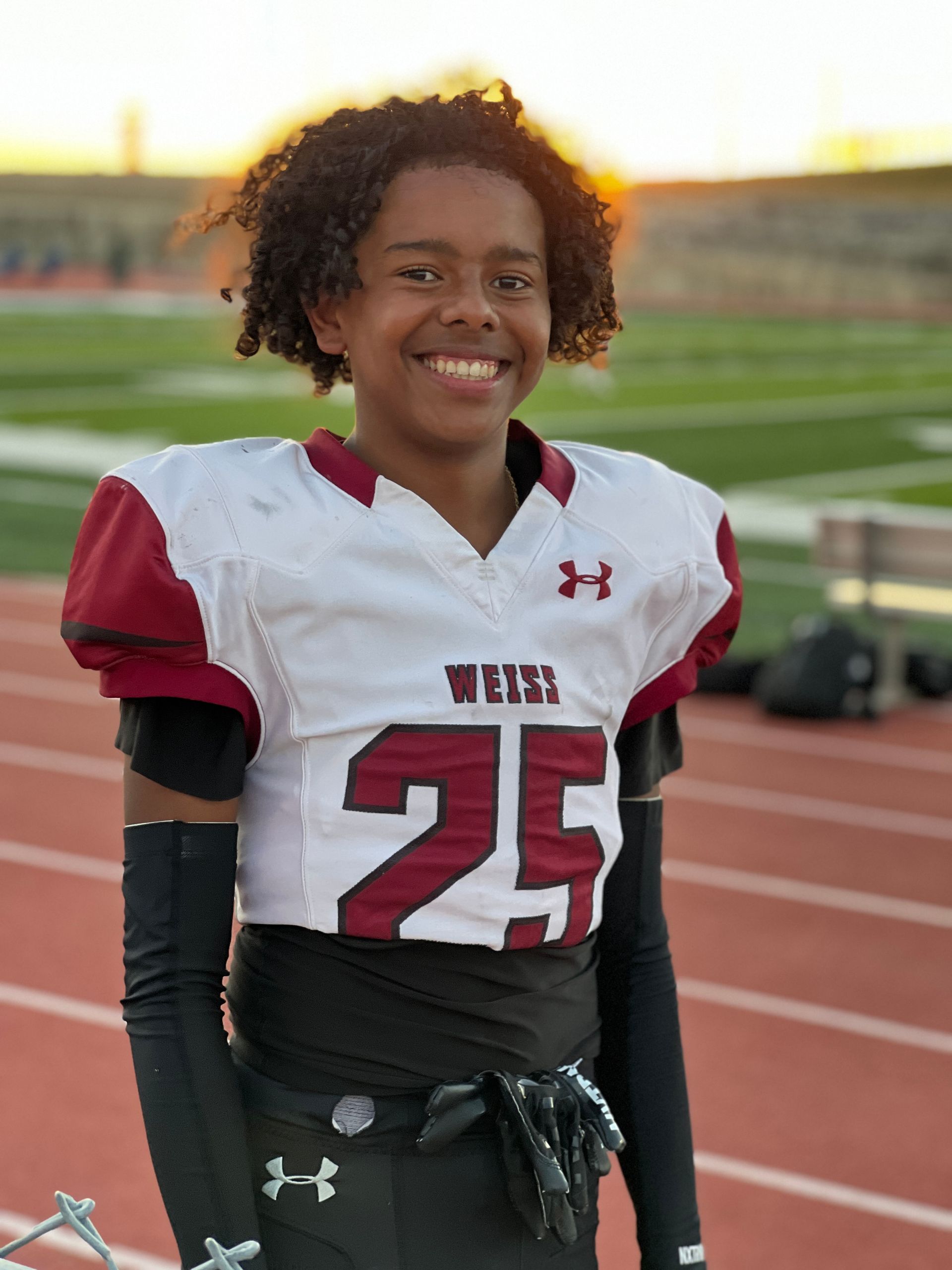 Young football player wearing a white and maroon jersey with the number 25 smiles on a track.