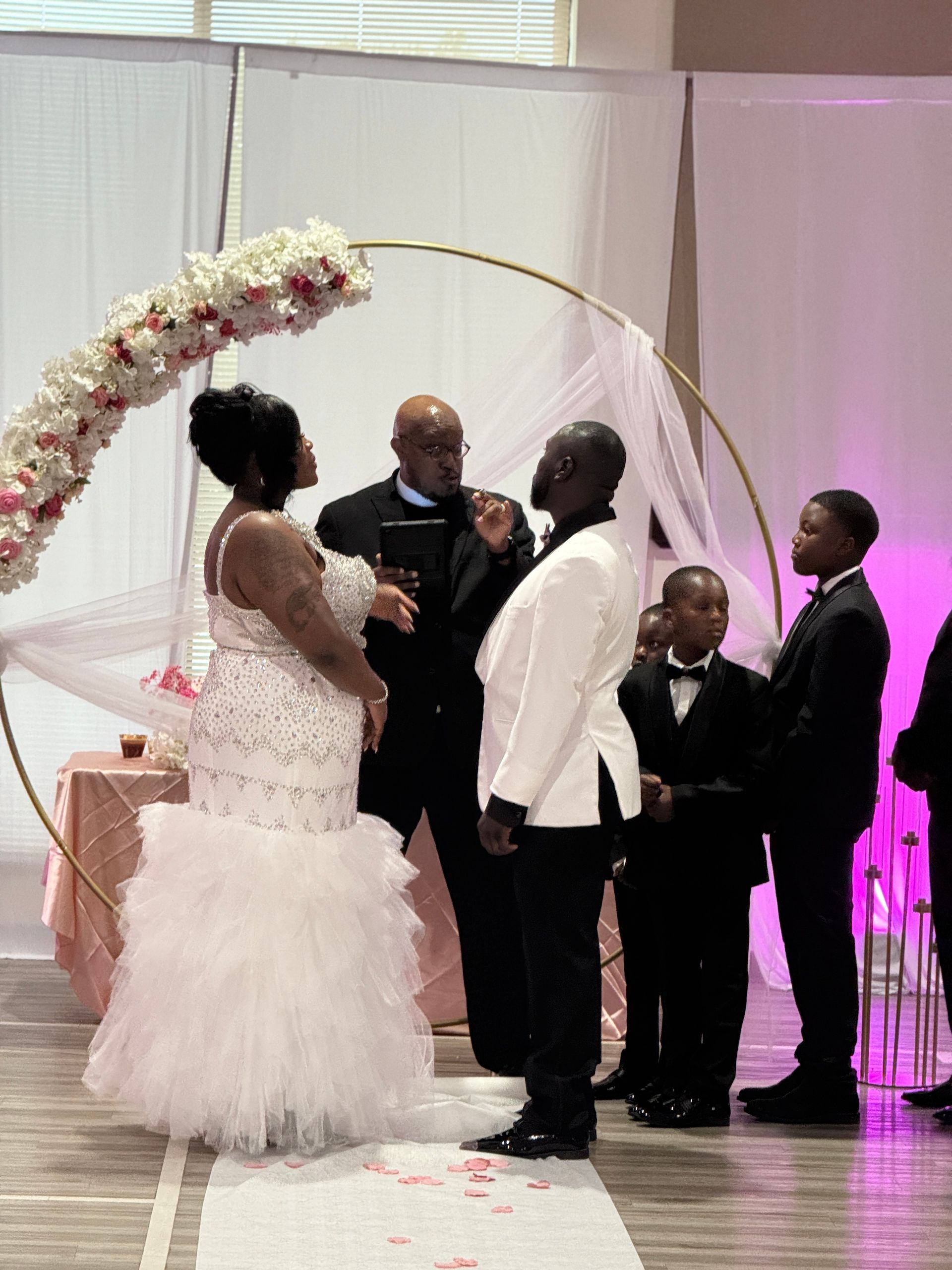 Wedding ceremony: Bride in white gown and groom in white jacket stand before officiant.  A floral arch frames the couple.