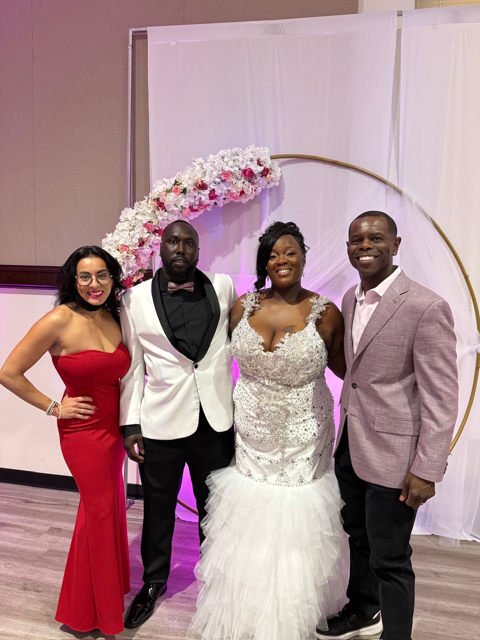 Four people pose for a photo at a wedding. A bride in a white gown and groom in a white tuxedo stand between two guests. They're in front of a floral arch.