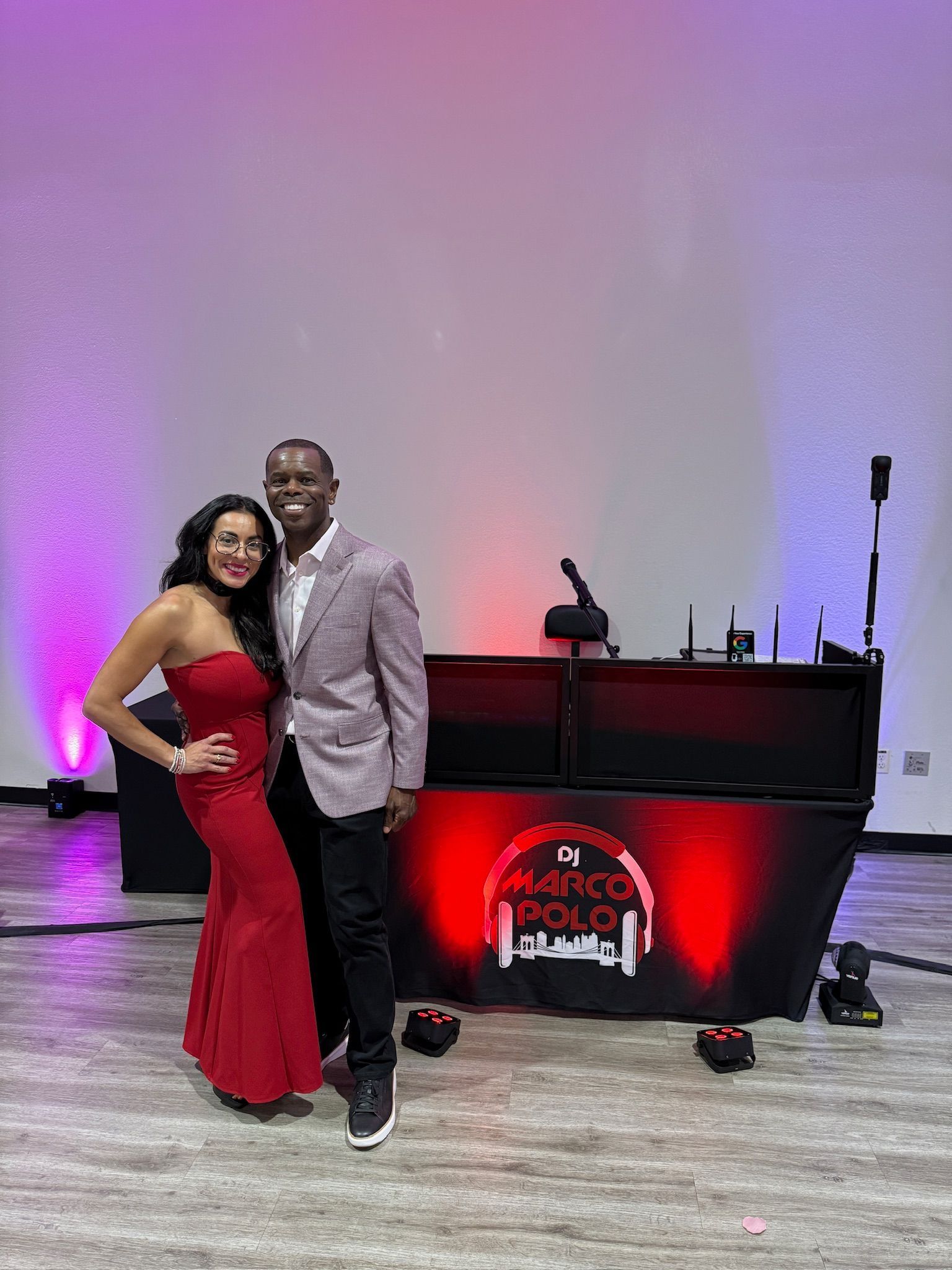 A man and woman pose in front of a DJ setup with purple and red lighting. The woman wears a red dress, and the man wears a grey blazer.