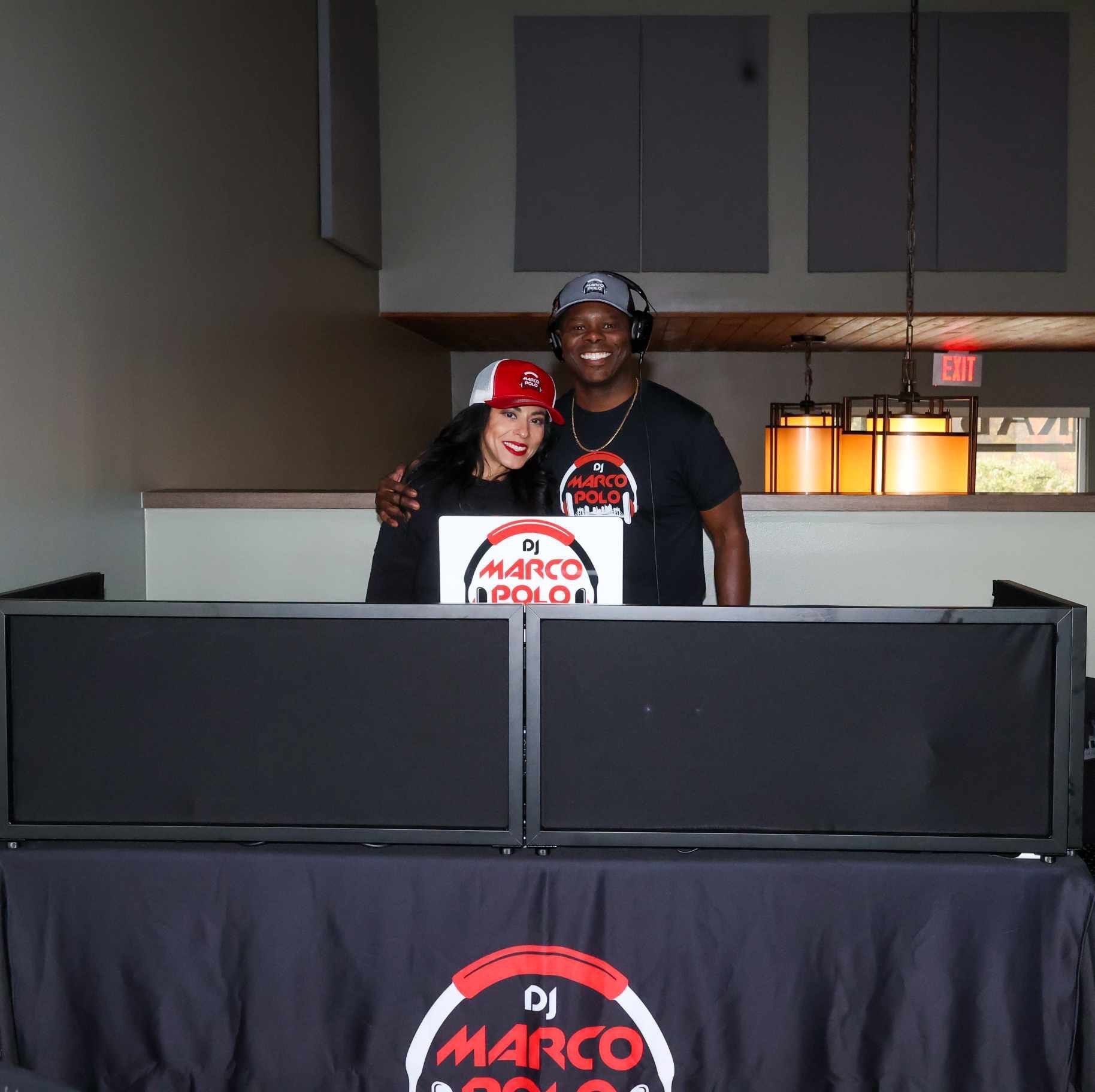 Two DJs, a man and a woman, smiling behind a DJ setup with a 