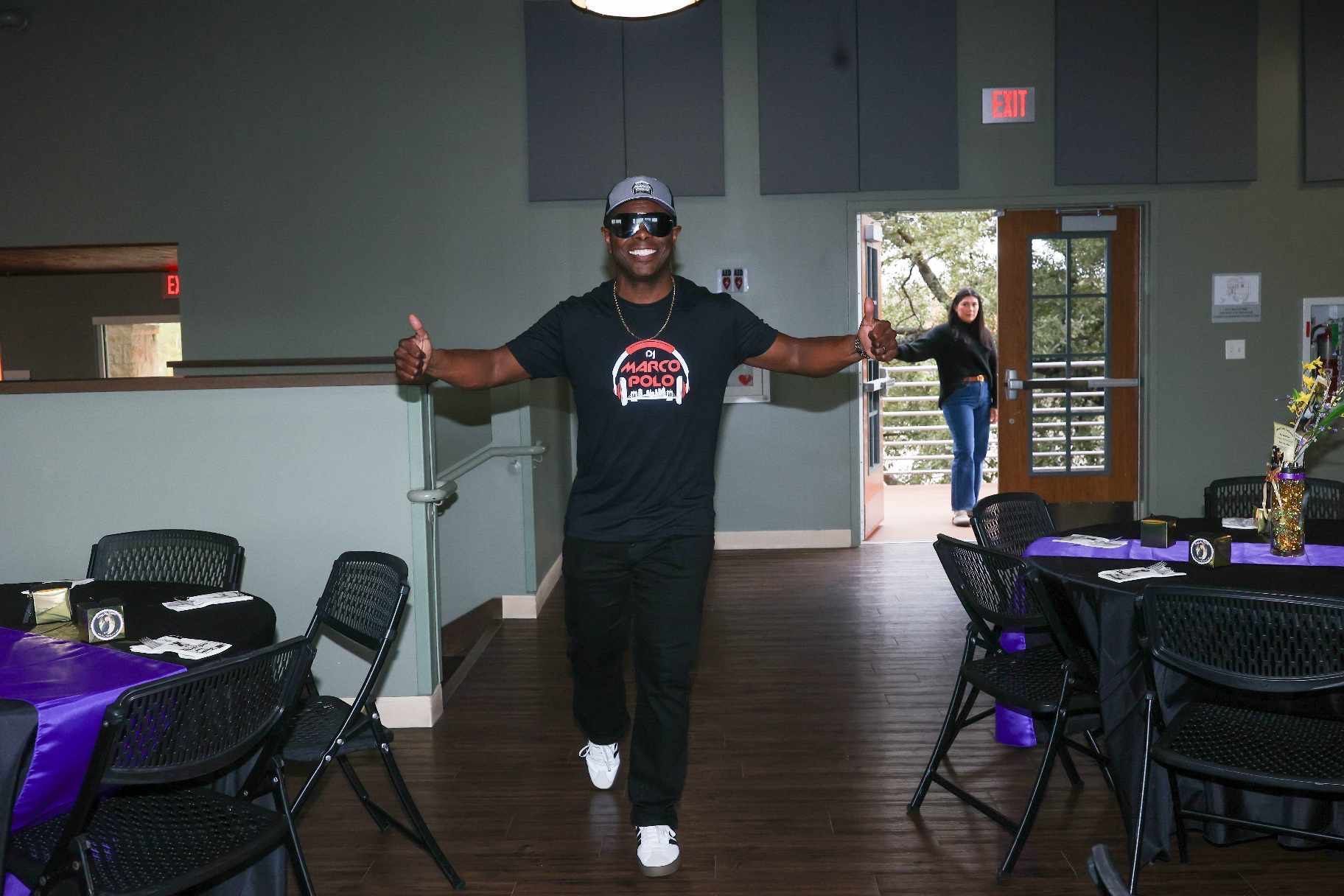 A man in sunglasses and a t-shirt strides toward the camera with his arms outstretched, entering a room with tables set for an event.