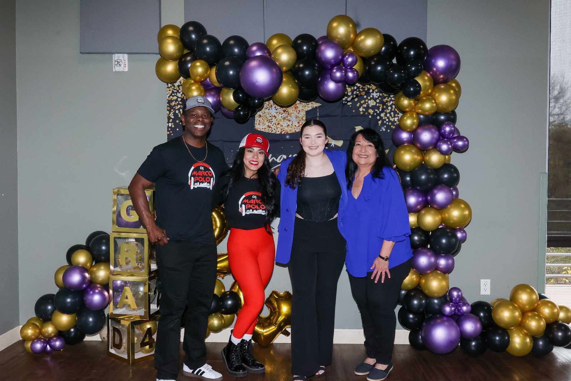 Four people pose in front of a balloon arch. Gold, black, and purple balloons frame a backdrop. The people smile and wear casual clothes.