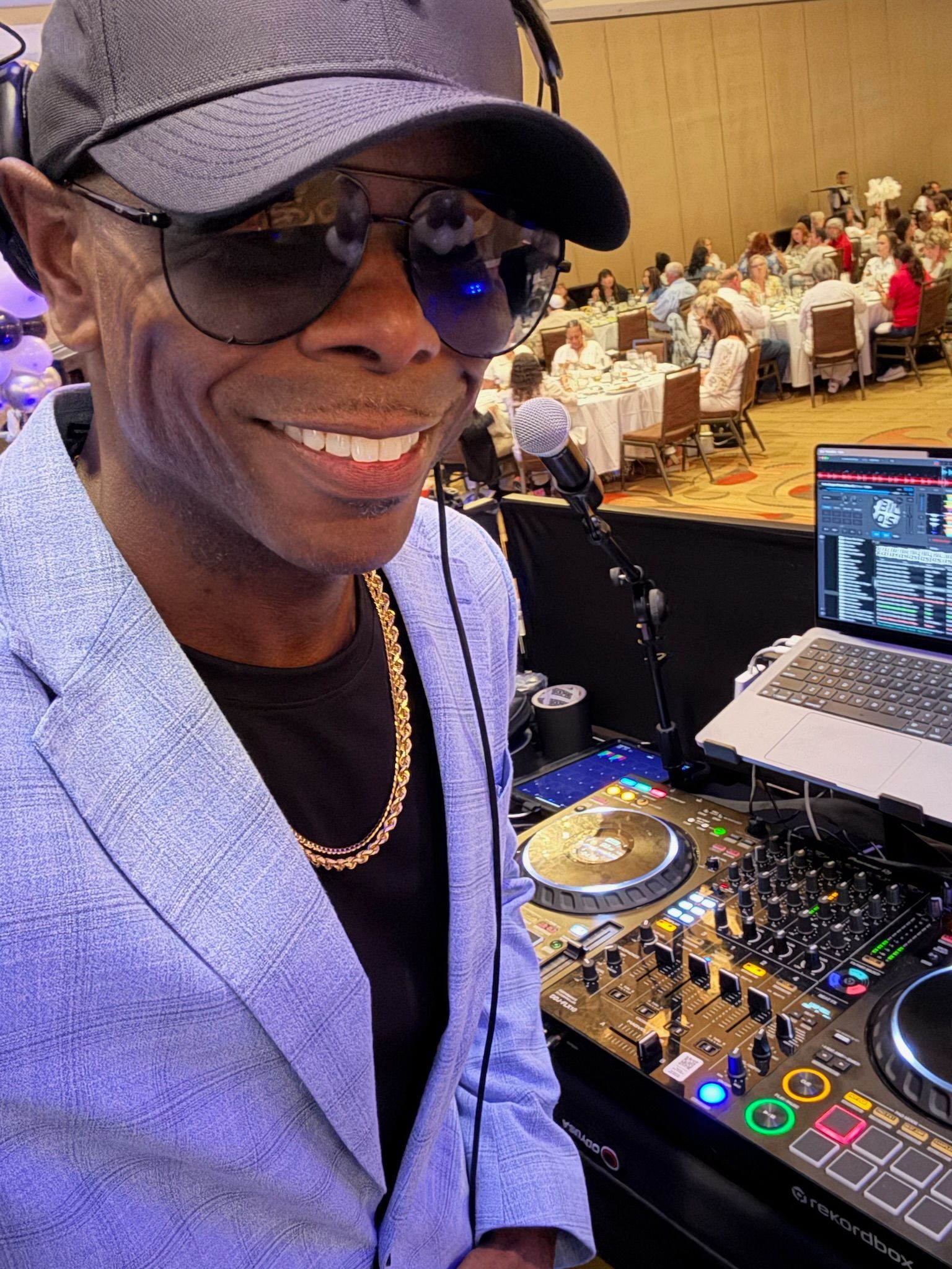 A DJ in a blazer and sunglasses smiles next to his equipment at a conference. In the background, people sit at tables.