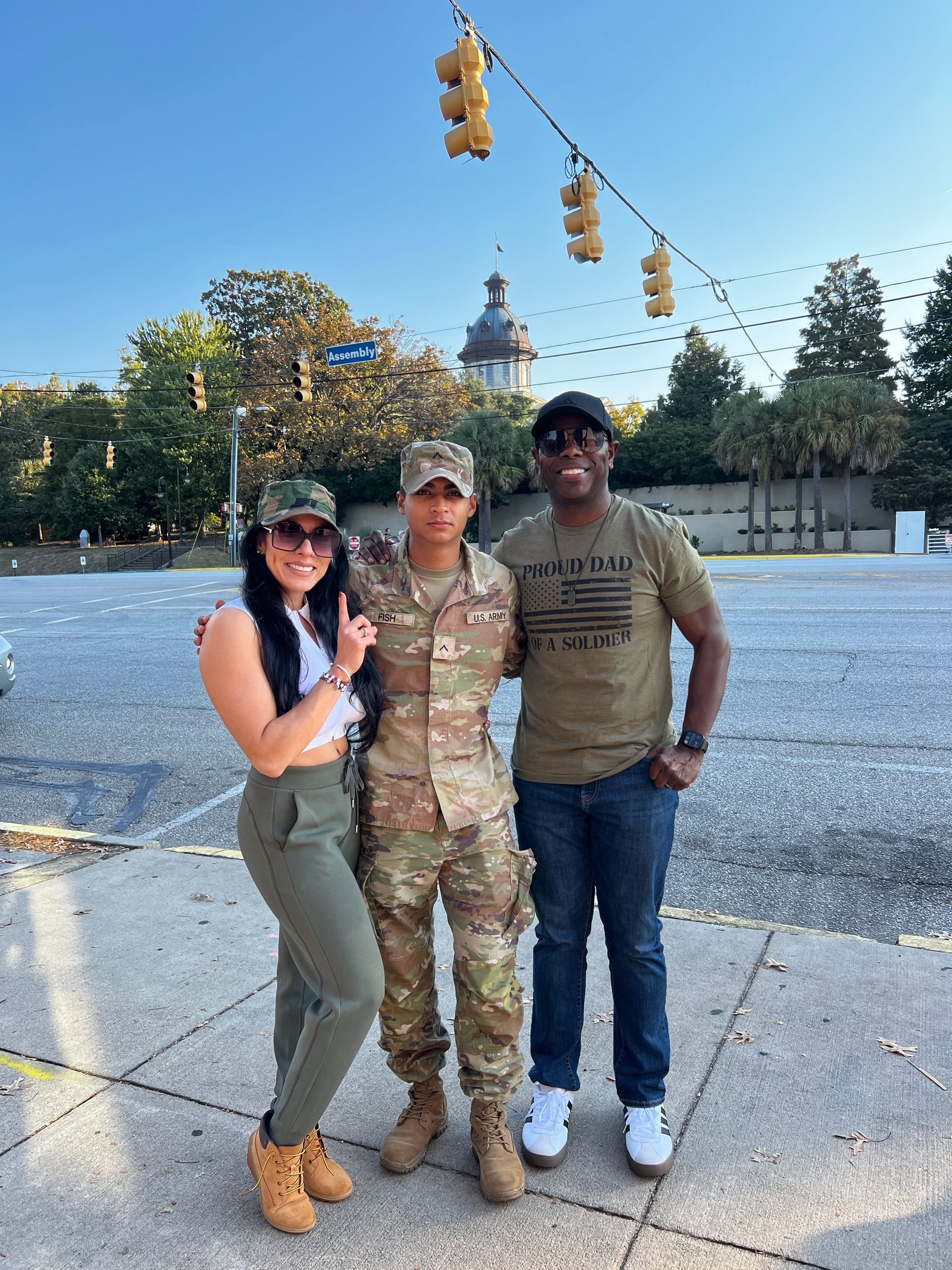Three people pose for a photo outdoors. A person in military uniform stands between two others in casual attire. The background includes a building and traffic lights.