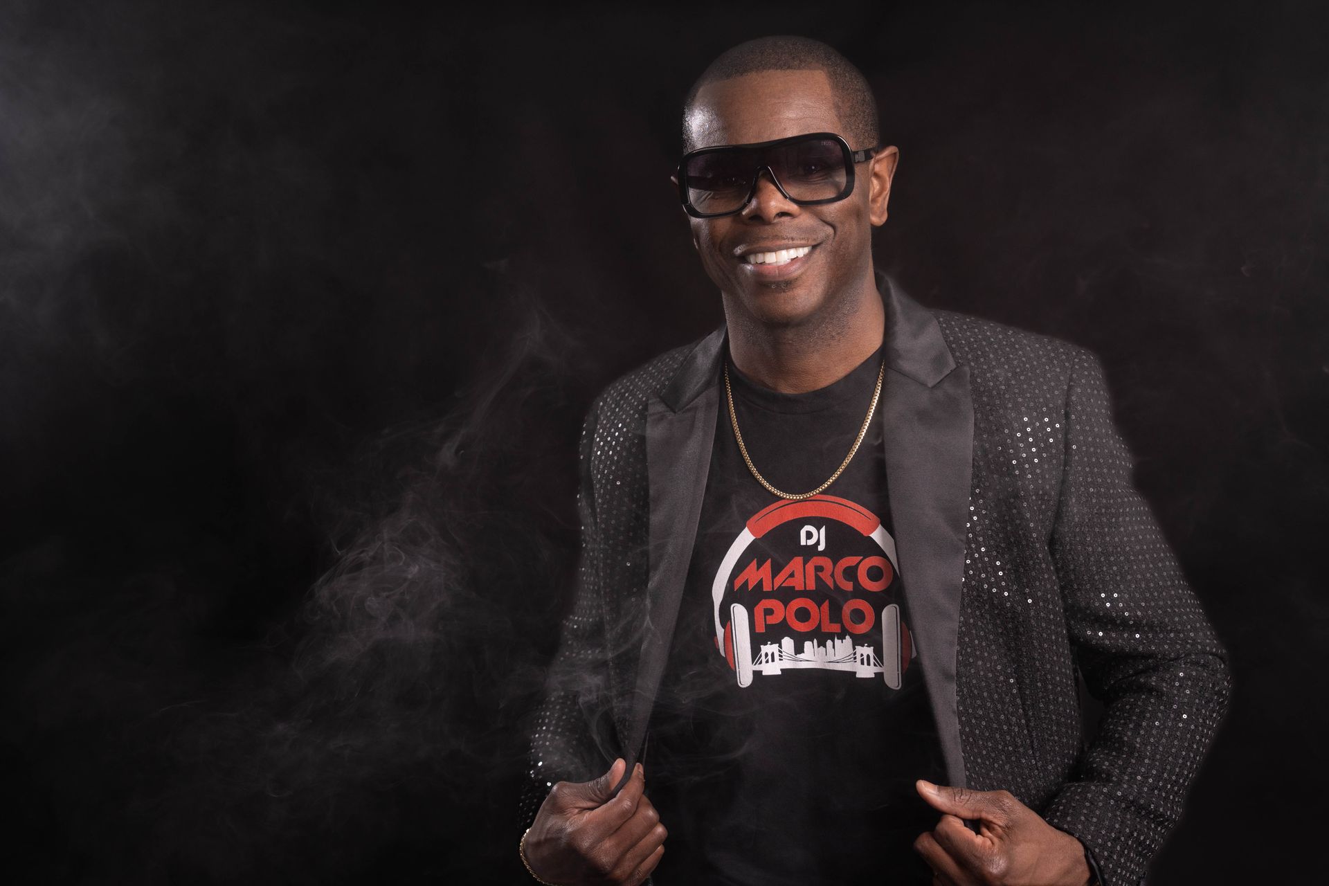 DJ Marco Polo in a dark setting, smiling, wearing sunglasses, a black t-shirt with his logo, and a sparkly jacket.