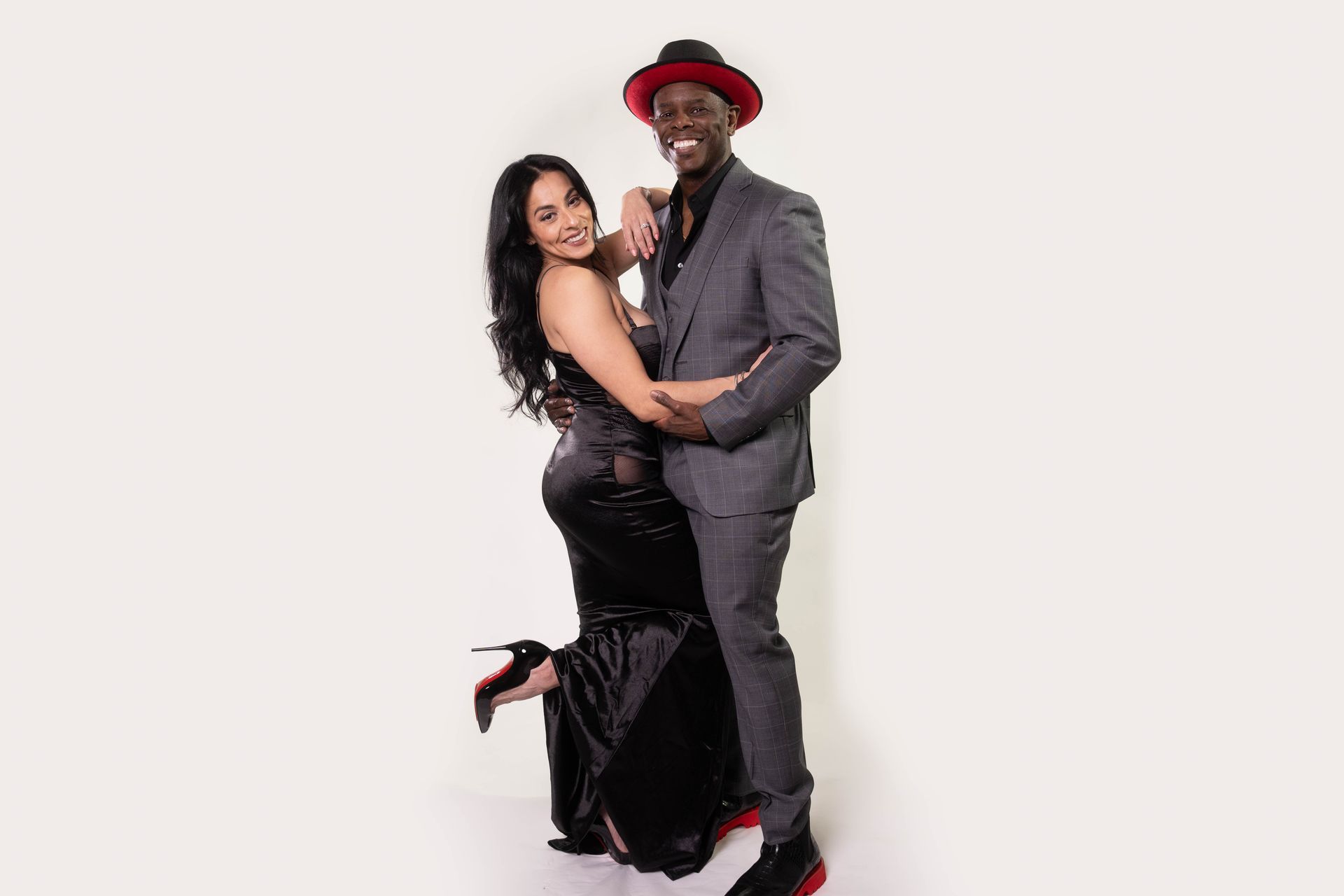 A smiling couple poses for a photo. The woman wears a black dress and the man a gray suit with a red-trimmed hat. They embrace.