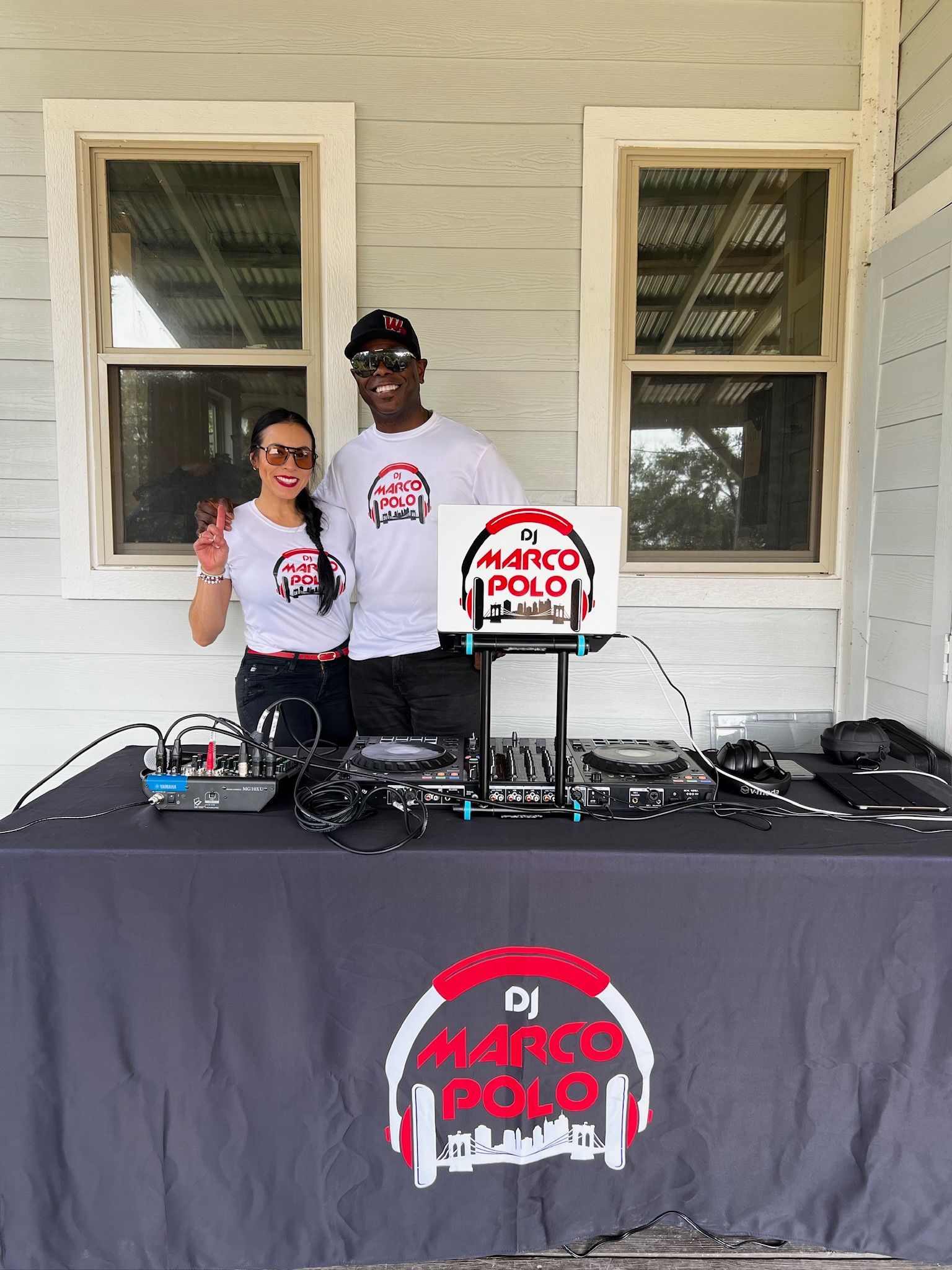 Two people, a man and woman, stand behind a DJ setup outside. They wear matching shirts with a logo and pose, one with a peace sign.
