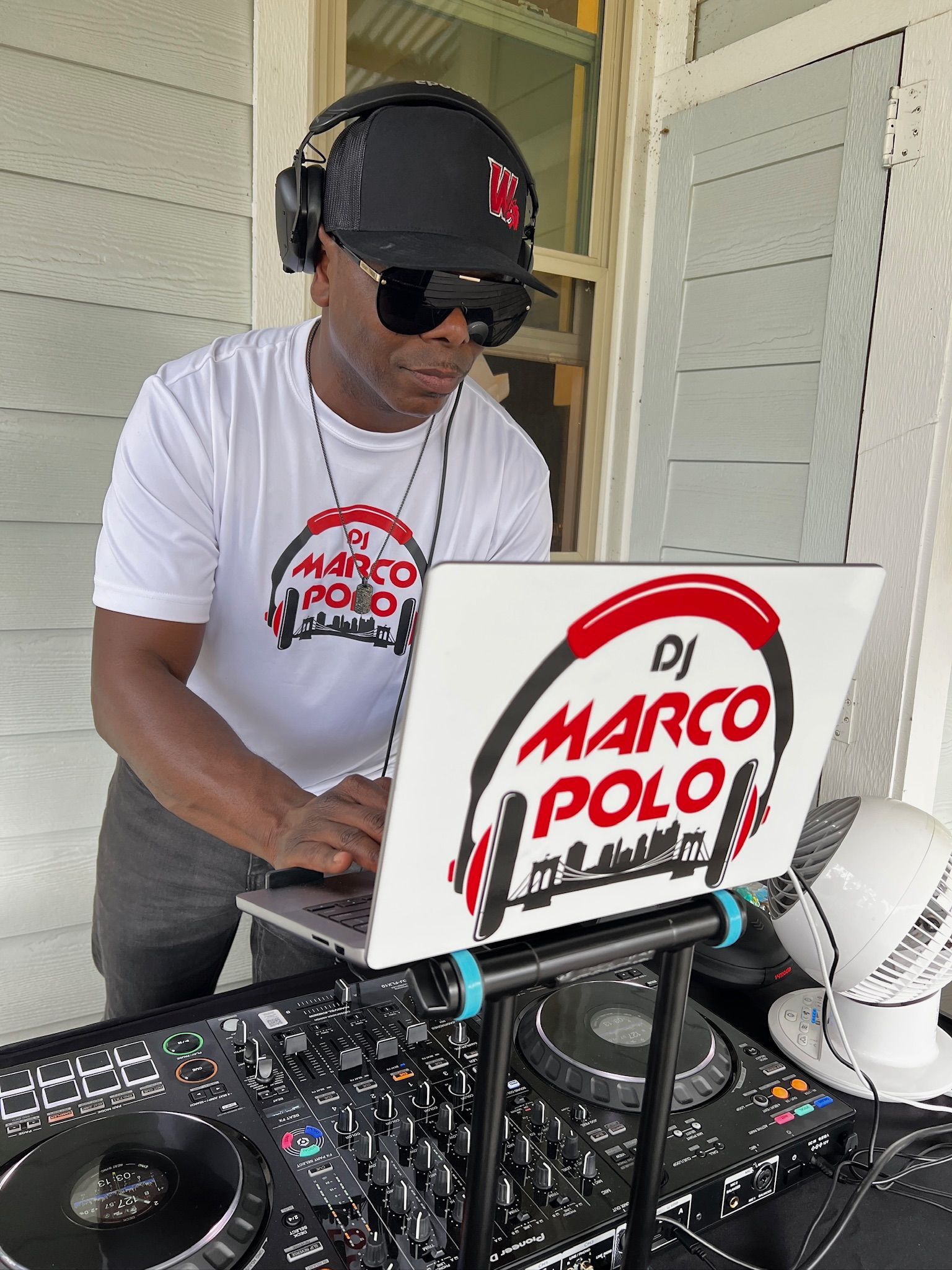 DJ Marco Polo stands at his equipment, wearing headphones and sunglasses. He is in a white t-shirt with his logo on the laptop.
