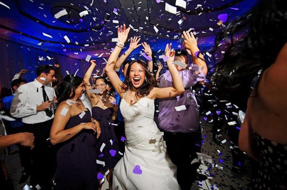 Bride dancing joyfully at a wedding reception, surrounded by friends, confetti, and blue lighting.