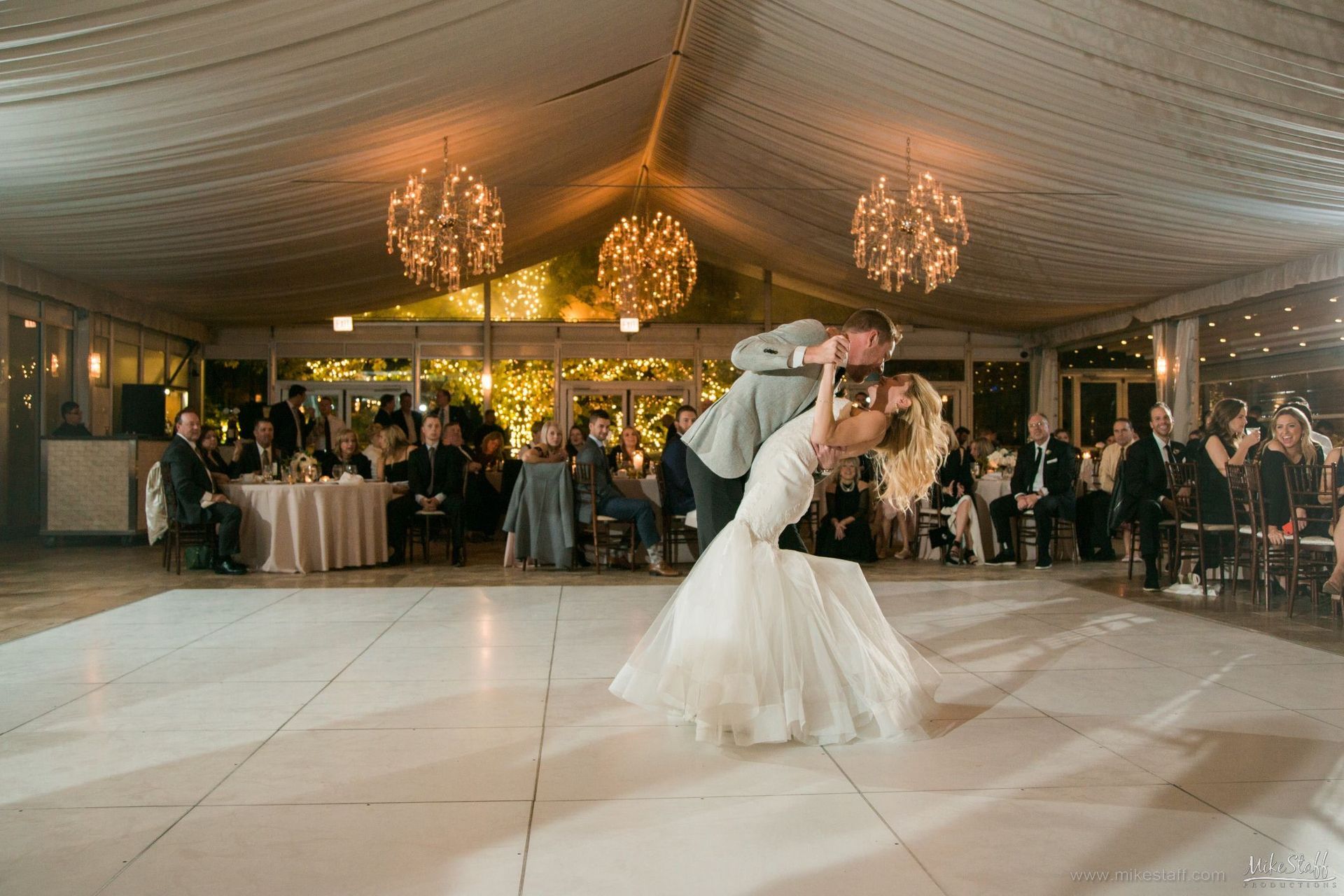 Bride and groom share a romantic dip during their first dance at a wedding reception. The reception is under a tent with overhead lights.