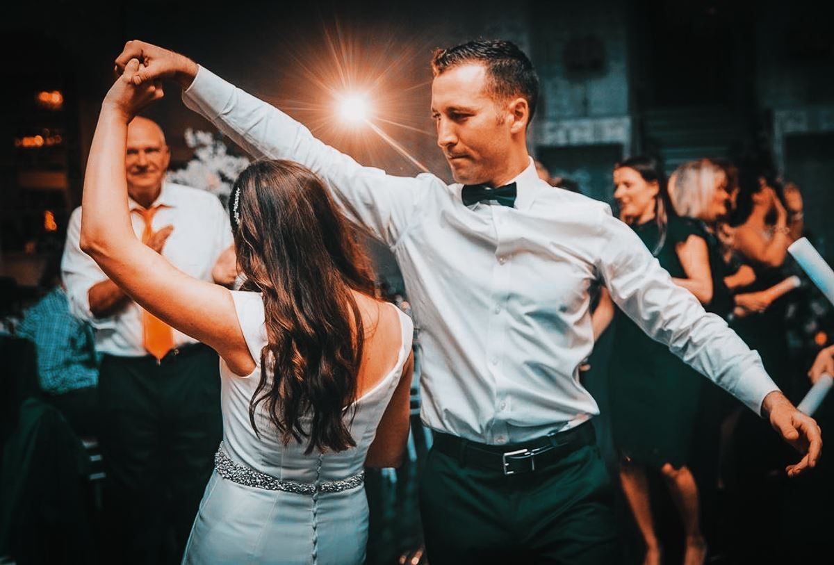 Couple dancing at a wedding reception; the man in a tuxedo, the woman in a white dress. Other guests are in the background.