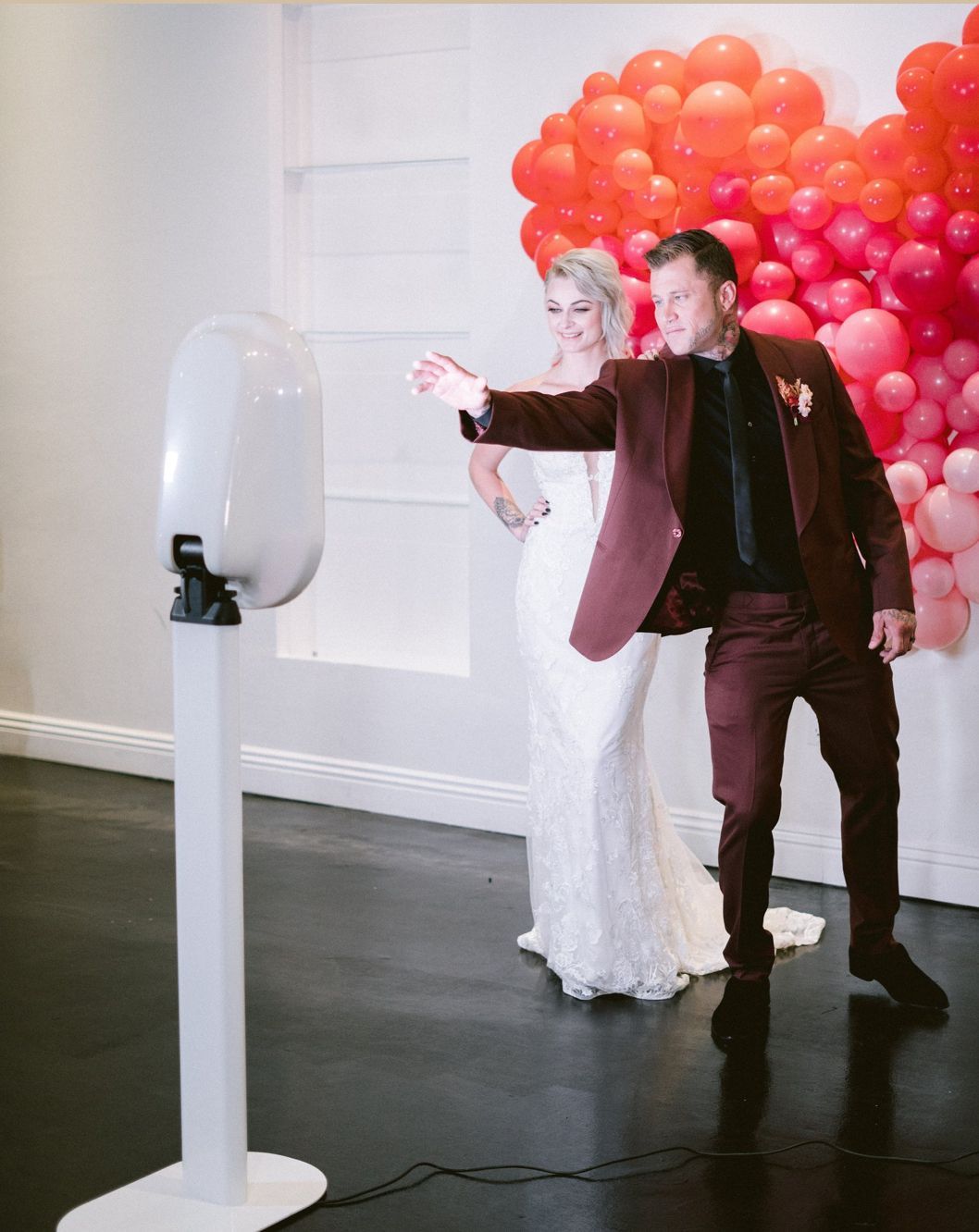 Newlyweds taking a photo in a photo booth; groom points towards camera, bride smiles, backdrop of heart-shaped balloons.