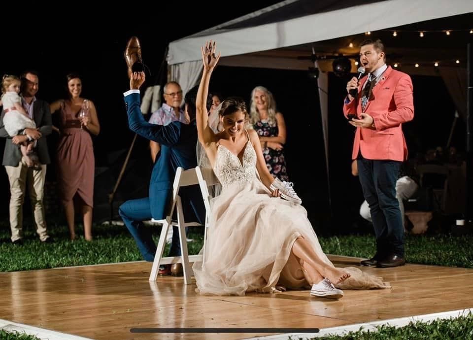 Bride and groom play a shoe game at their outdoor wedding reception. The bride has her arm raised, smiling, while the groom holds up his shoe.