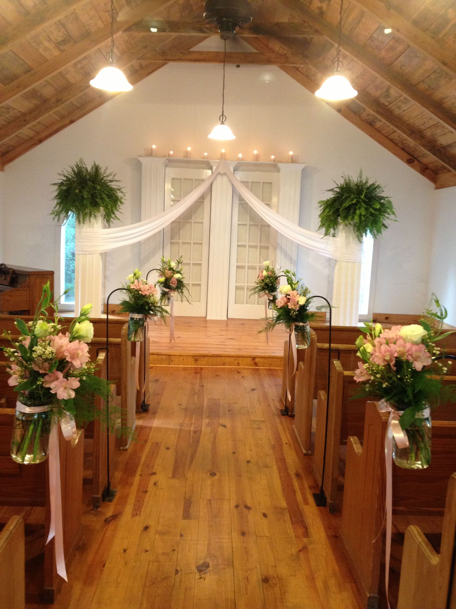 Inside a small chapel decorated for a wedding. Wooden floor and pews, flowers in mason jars, sheer white curtains, and two floral arrangements at the back doors.