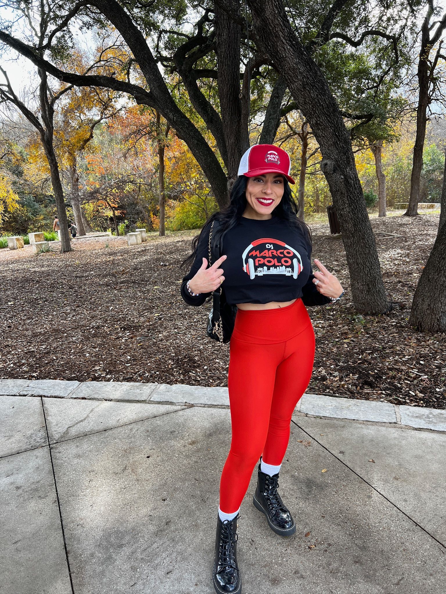 Woman in red leggings and a baseball cap points at her black crop top with text, standing outside on a path in front of trees.