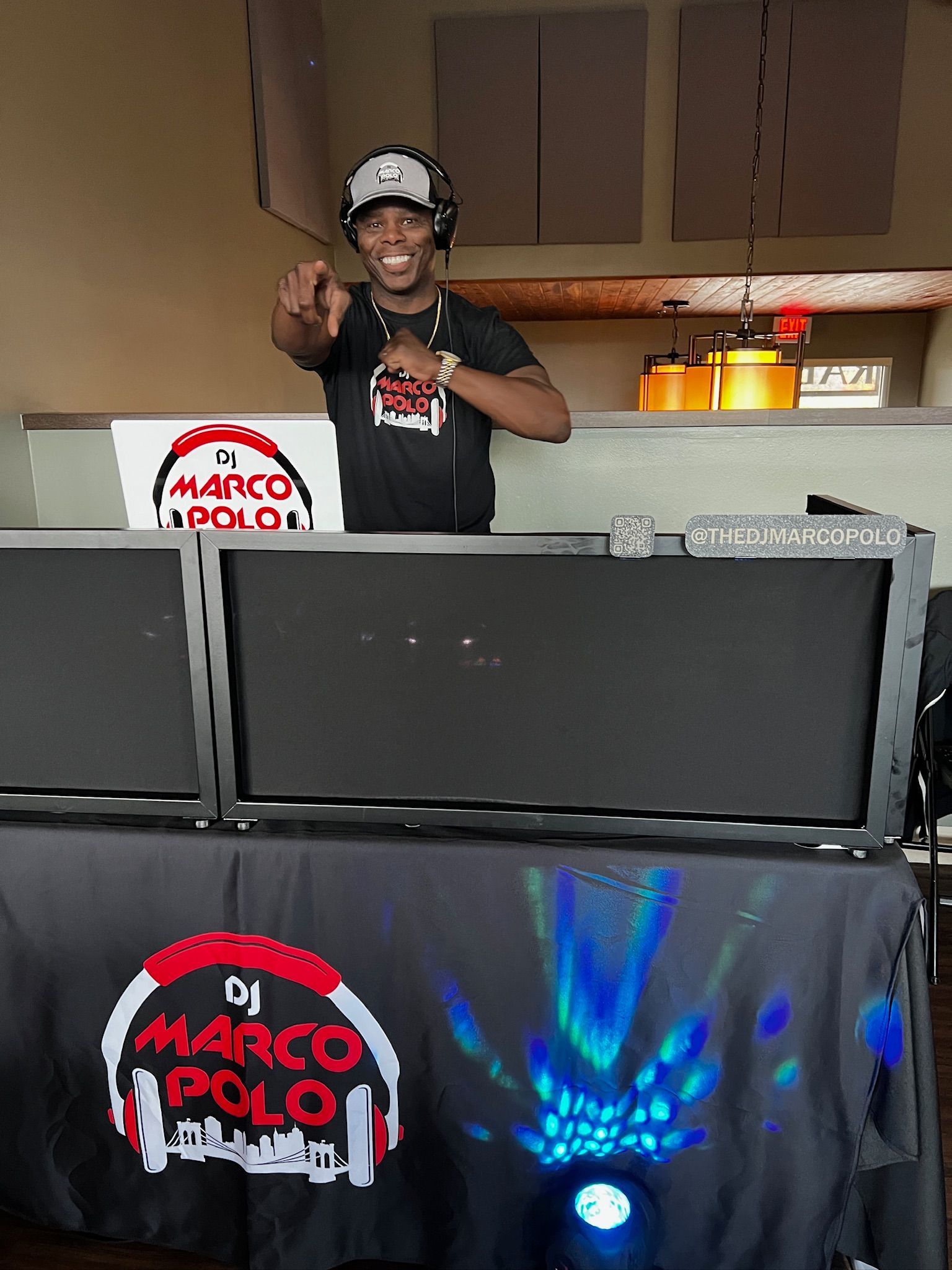 DJ Marco Polo at a party, pointing at the viewer with a smile. He is behind a table with DJ equipment, a logo on the front.