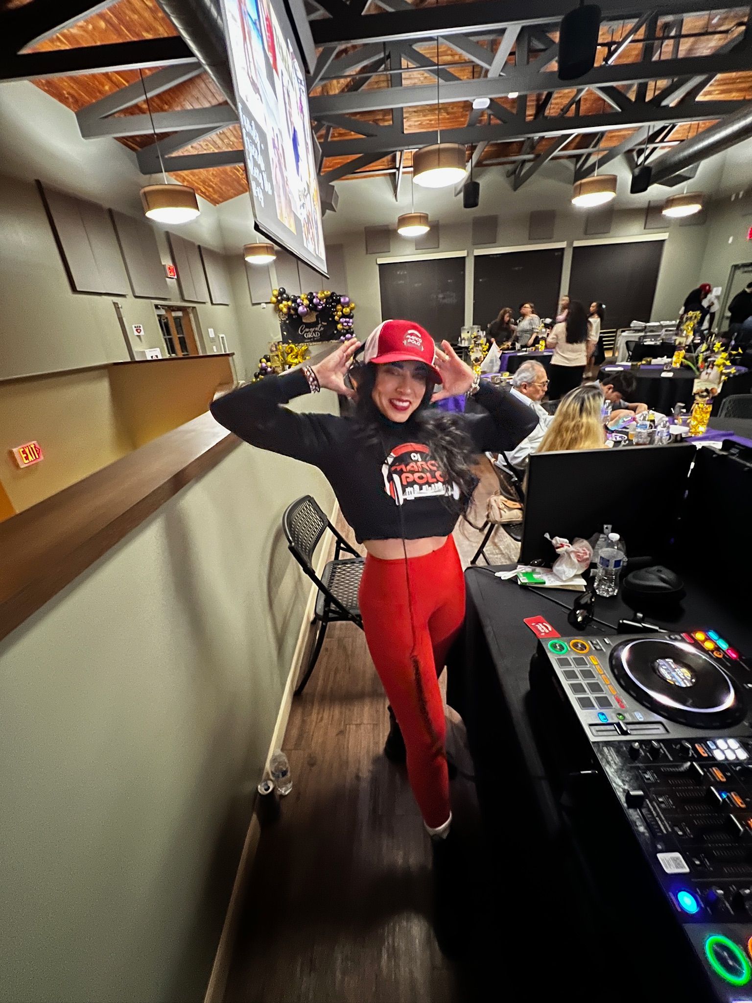 A woman in a red hat and pants smiles, raising her hands near her head while posing next to a DJ setup.
