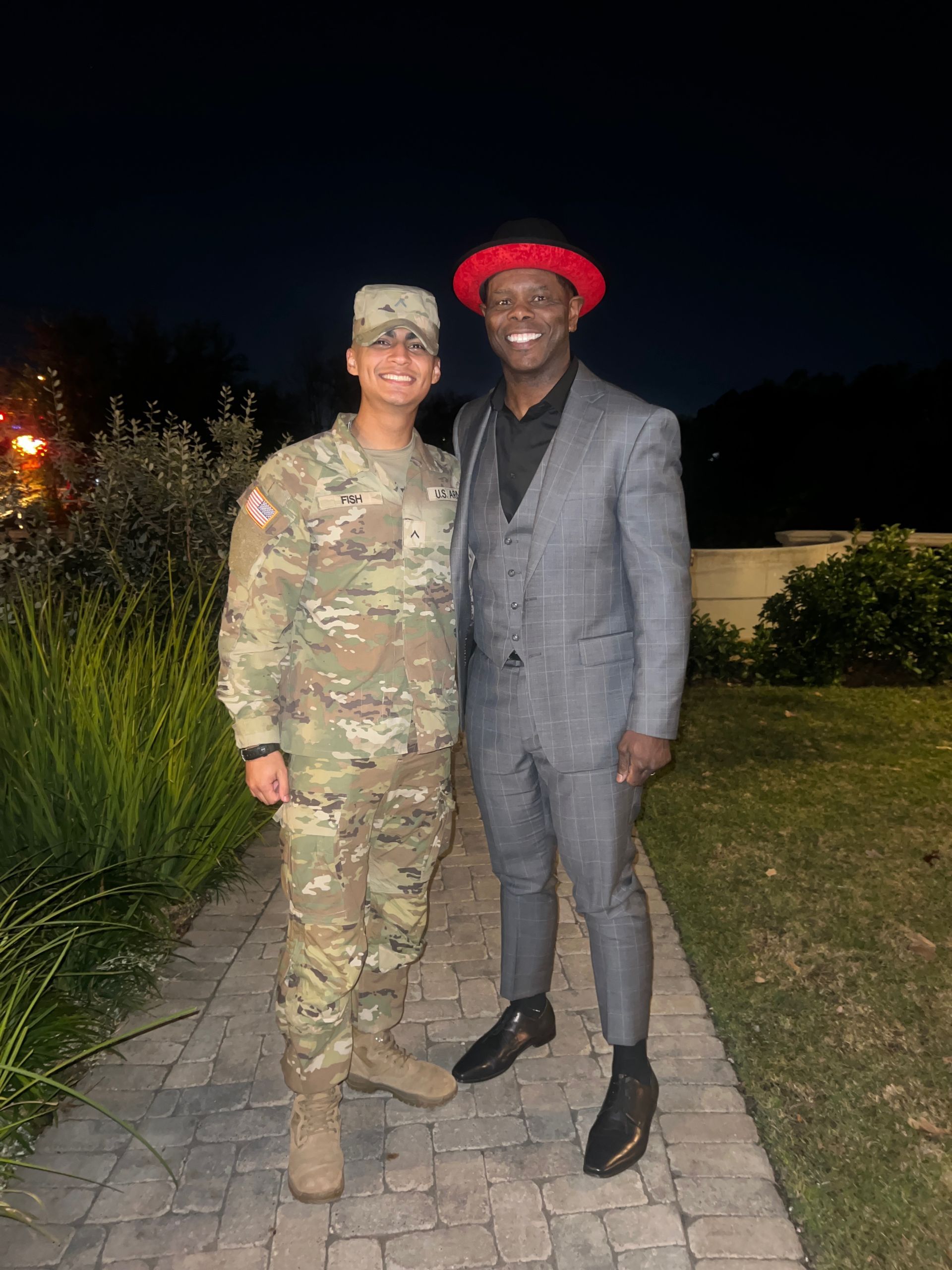 Soldier in uniform poses with a man in a grey suit, a red hat, smiling, at night on a brick walkway.