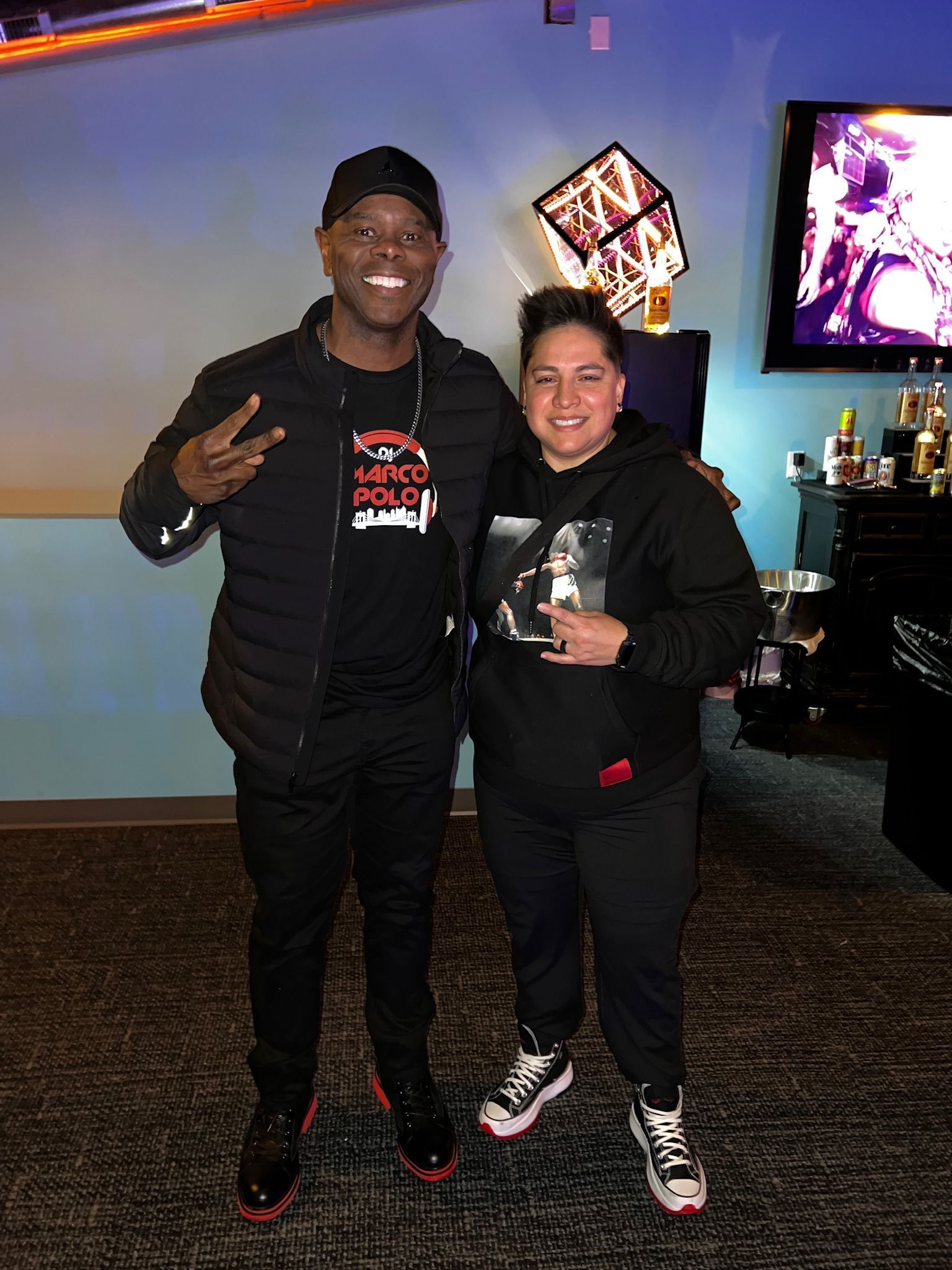 Two people pose for a photo in front of a blue wall. A man in black makes a peace sign. A person wearing a hoodie and sneakers stands beside him.