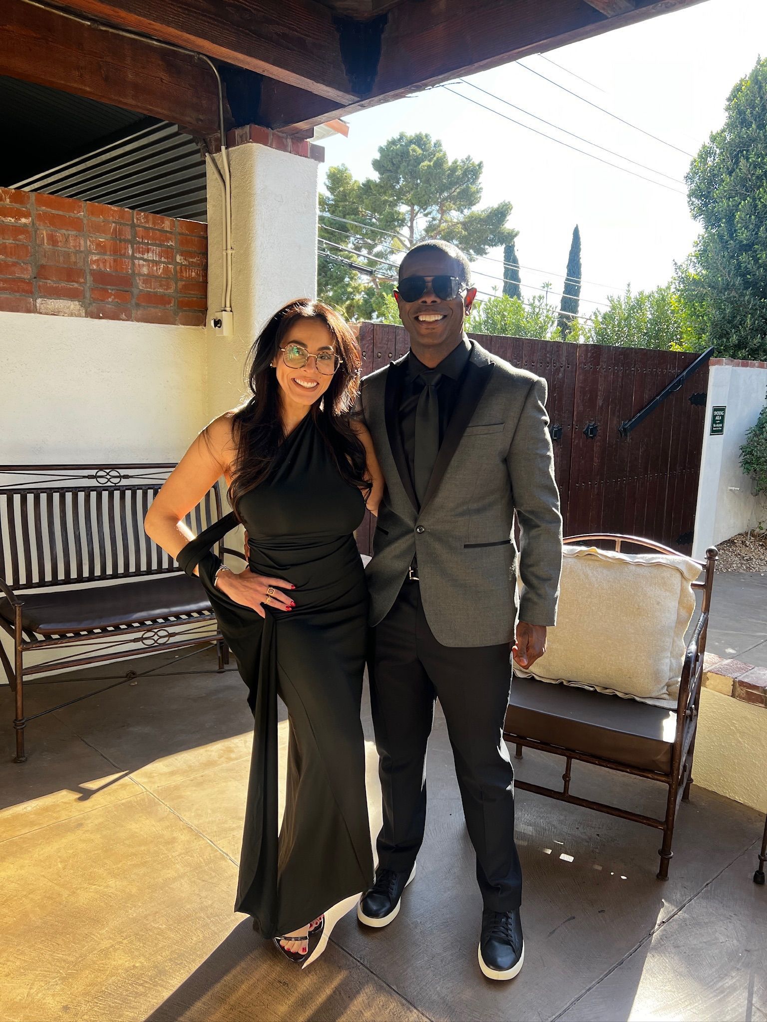 Woman in black gown and man in gray suit pose outside. The sun is shining.