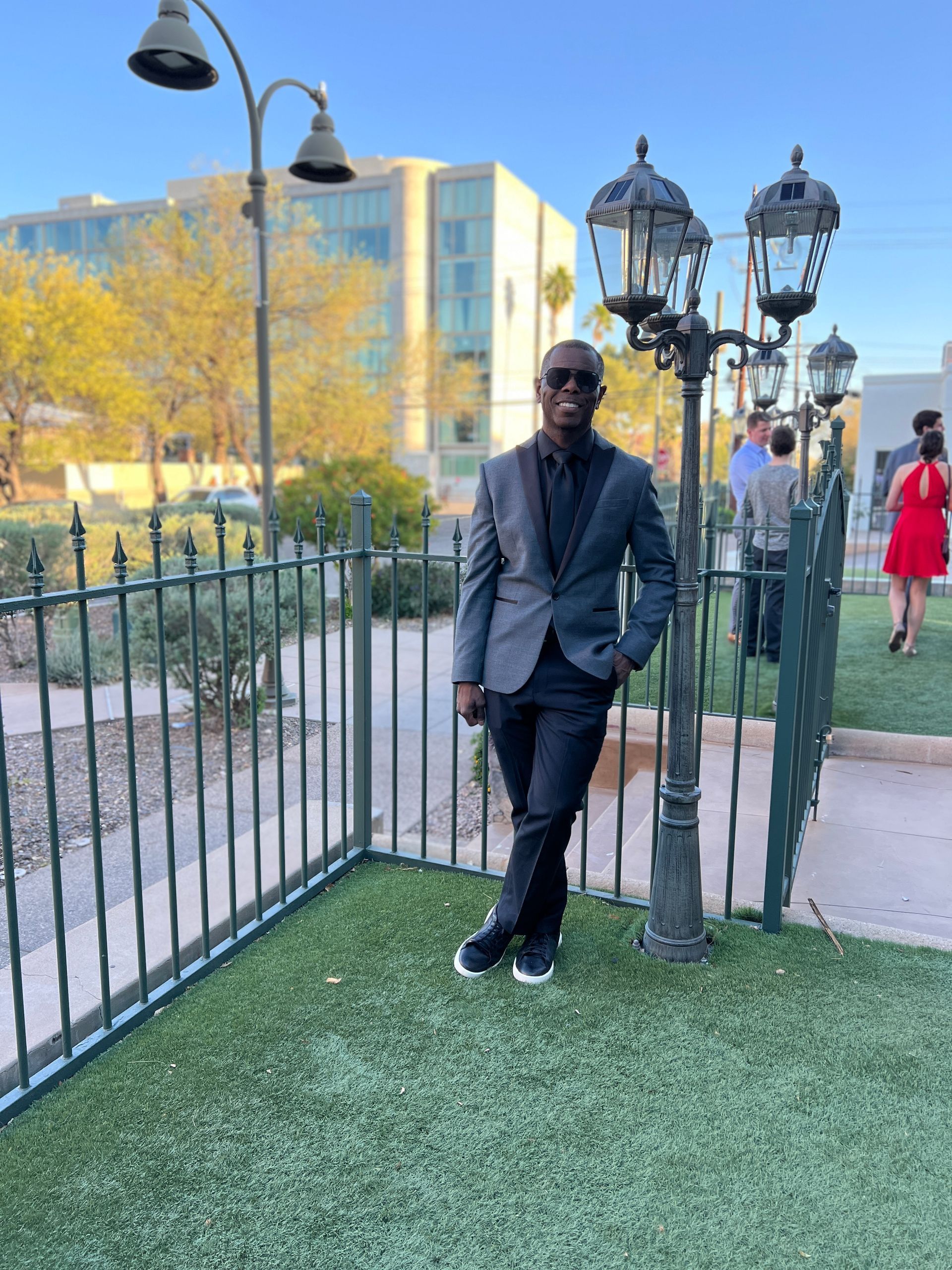 Man in a gray suit leans against a lamp post in a grassy area. Buildings and a wrought-iron fence are in the background.