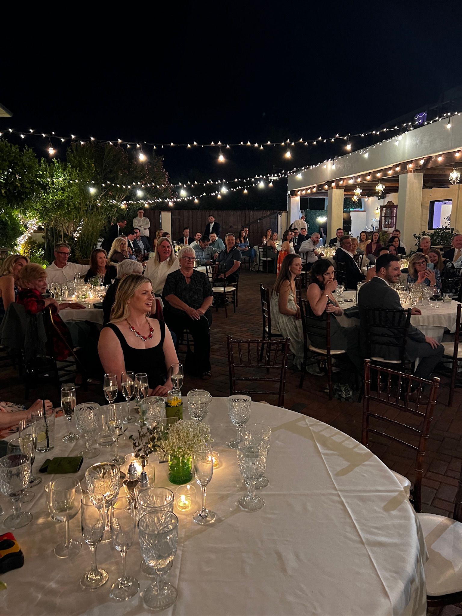 Outdoor nighttime gathering with people seated at tables, string lights overhead.