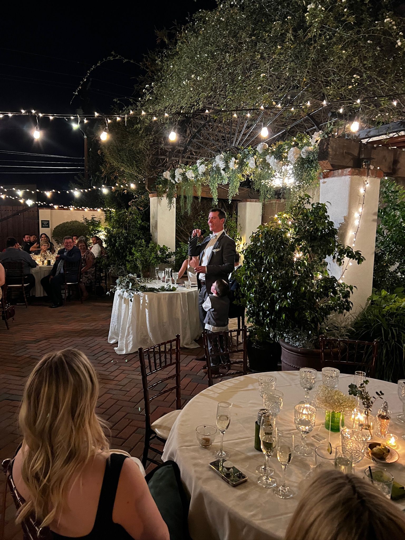 Man giving a speech at an outdoor wedding reception. Guests are seated at tables, and string lights illuminate the venue.