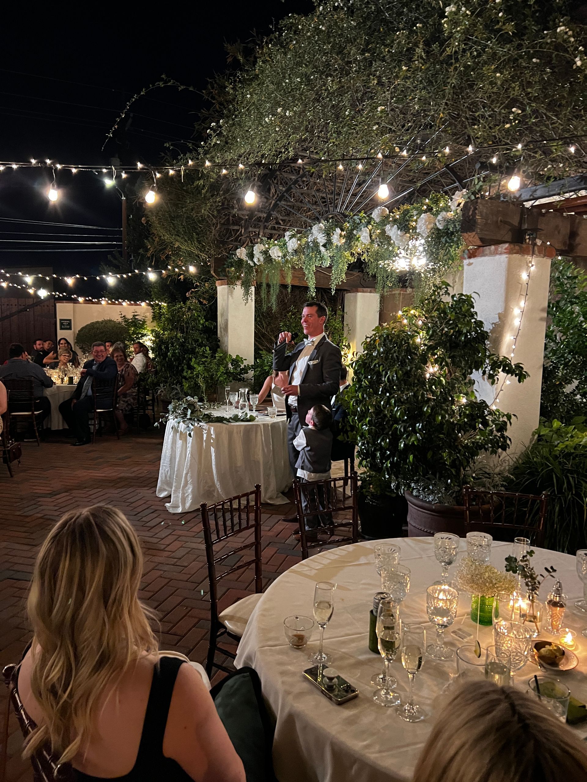 Man giving a speech at an outdoor wedding reception at night. Guests seated at tables with candles and string lights overhead.
