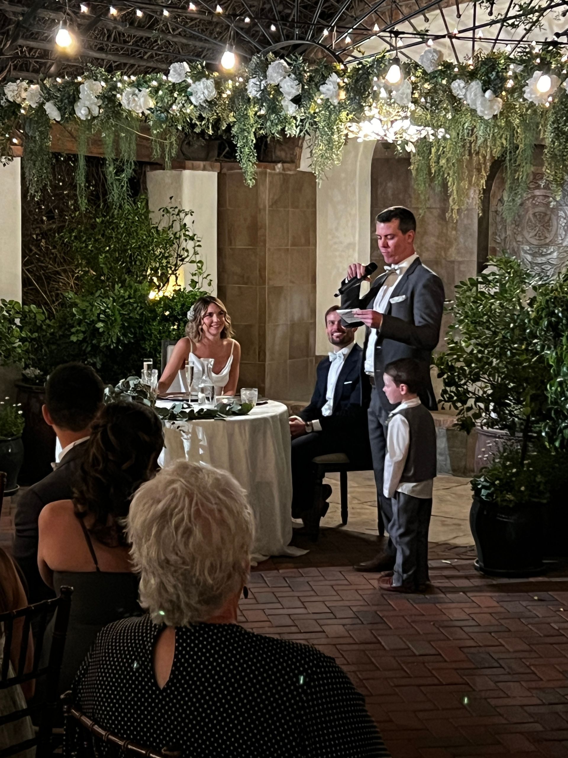 Man giving a speech at a wedding reception. Bride and groom seated at a table, decorated with flowers and greenery. Guests are seated.