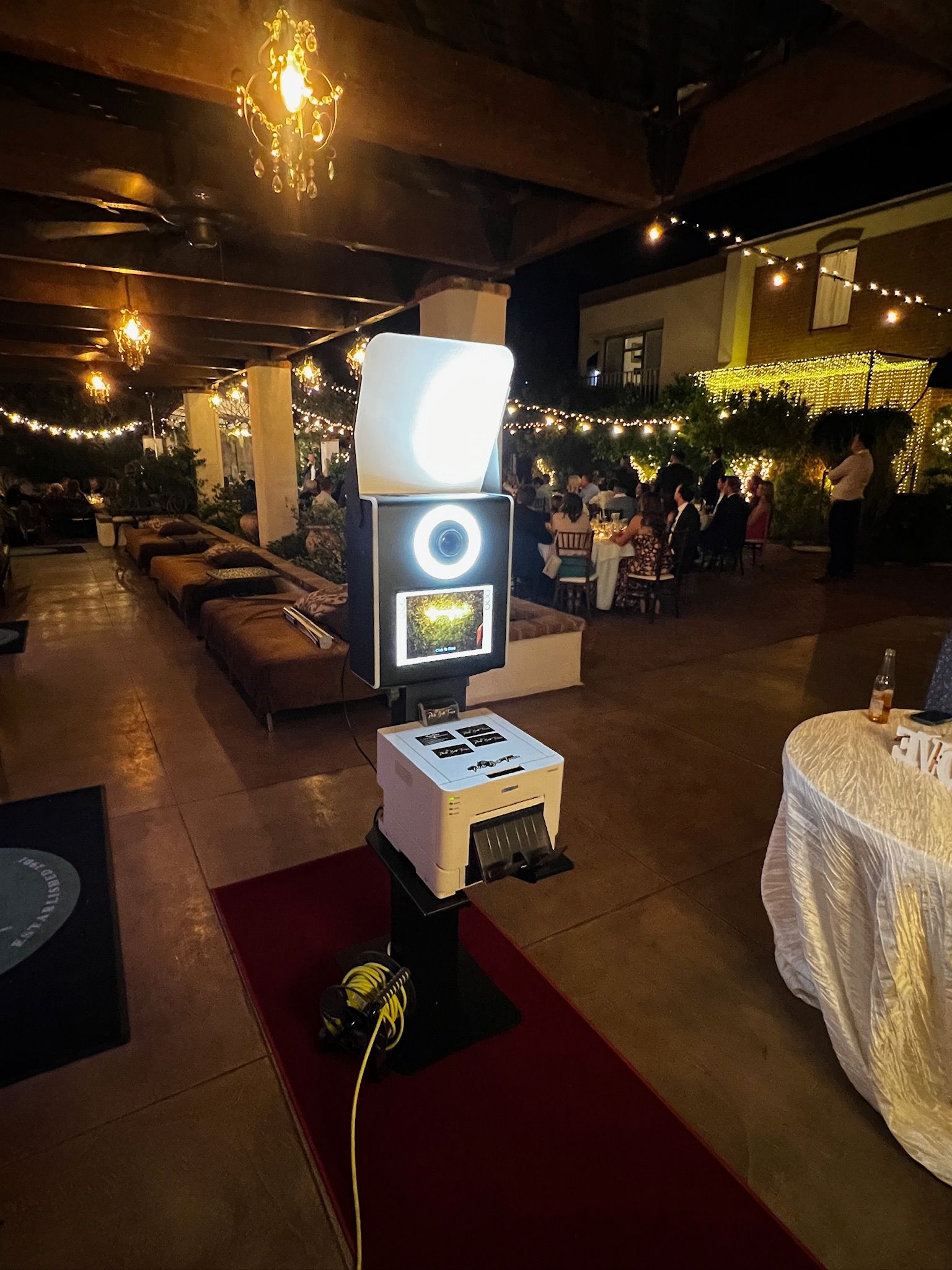Photo booth set up at an outdoor event. The booth has a white light, screen, and printer on a red carpet, with guests and lights in the background.
