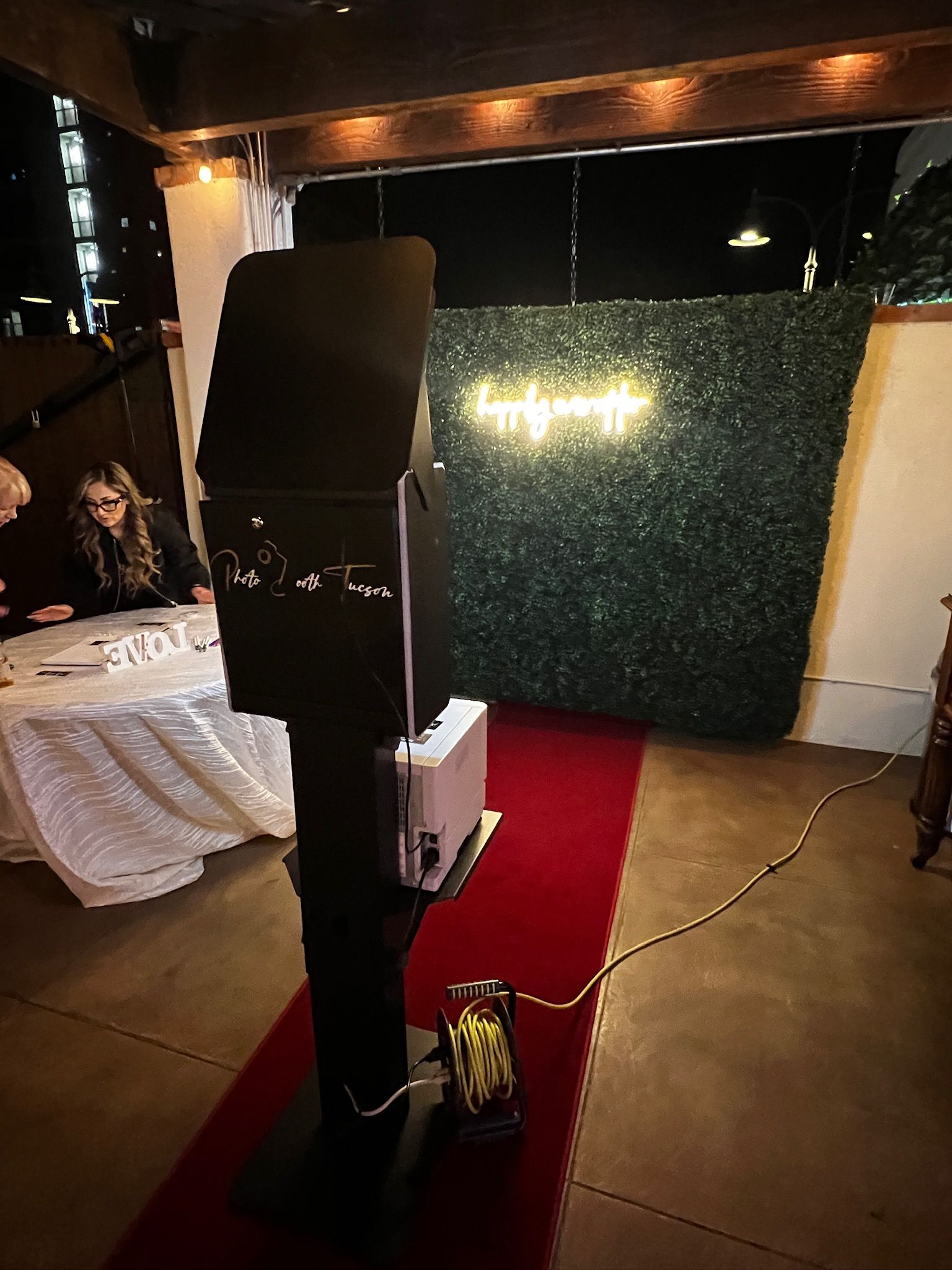 A photo booth with a red carpet and a green backdrop at an outdoor event. A woman sits at a table in the background.
