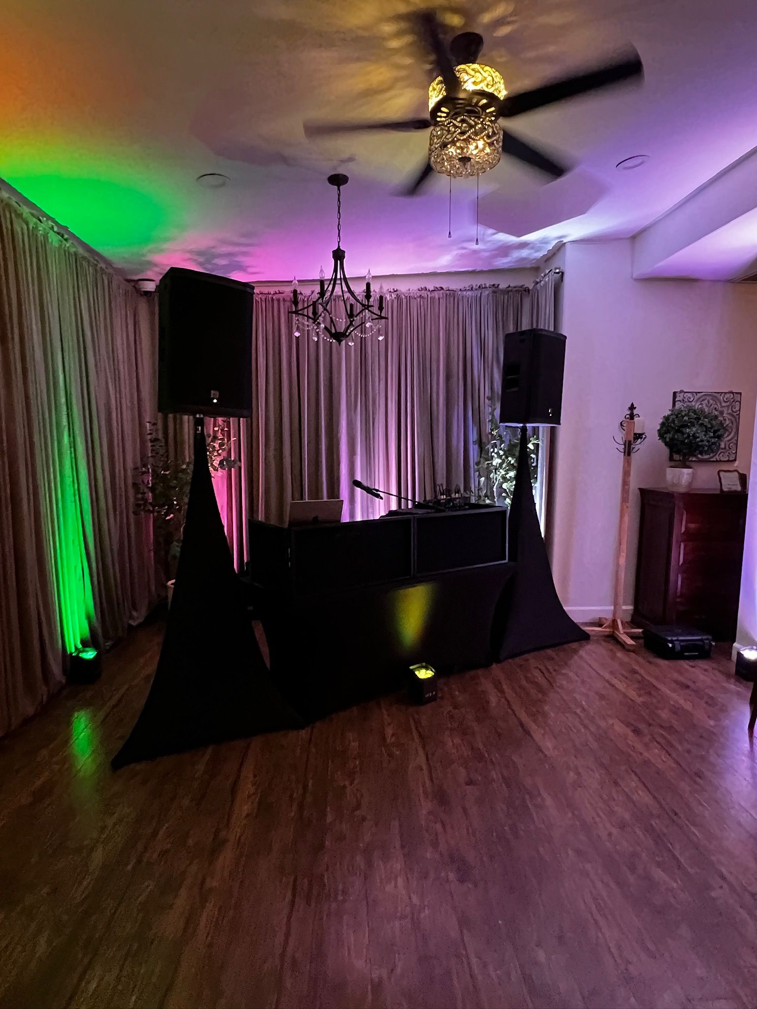 DJ setup in a room with colorful lighting, speakers, a table, and a chandelier. The floor is hardwood.