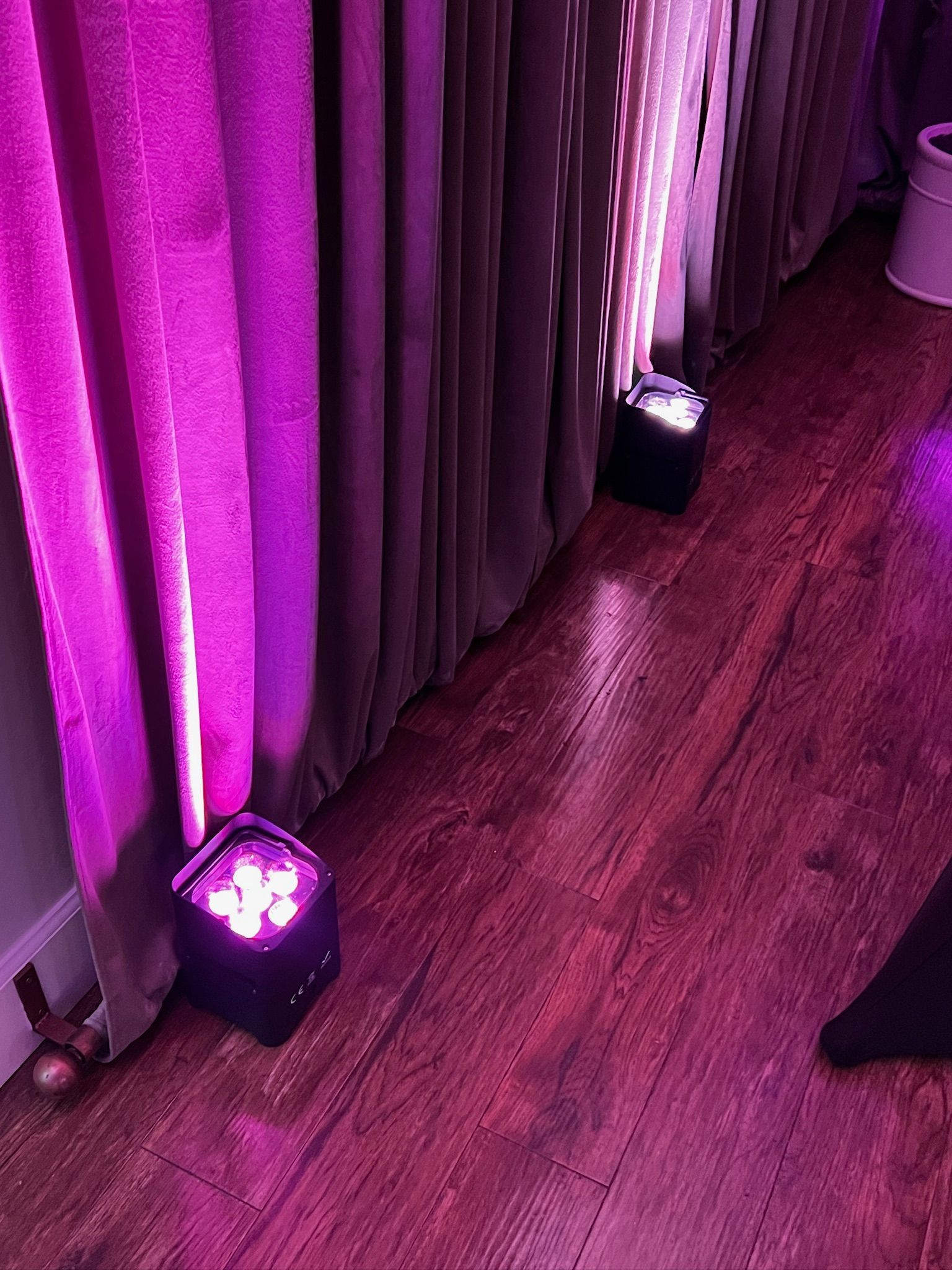 Two purple stage lights illuminate curtains and hardwood flooring. One light is next to a pink curtain.