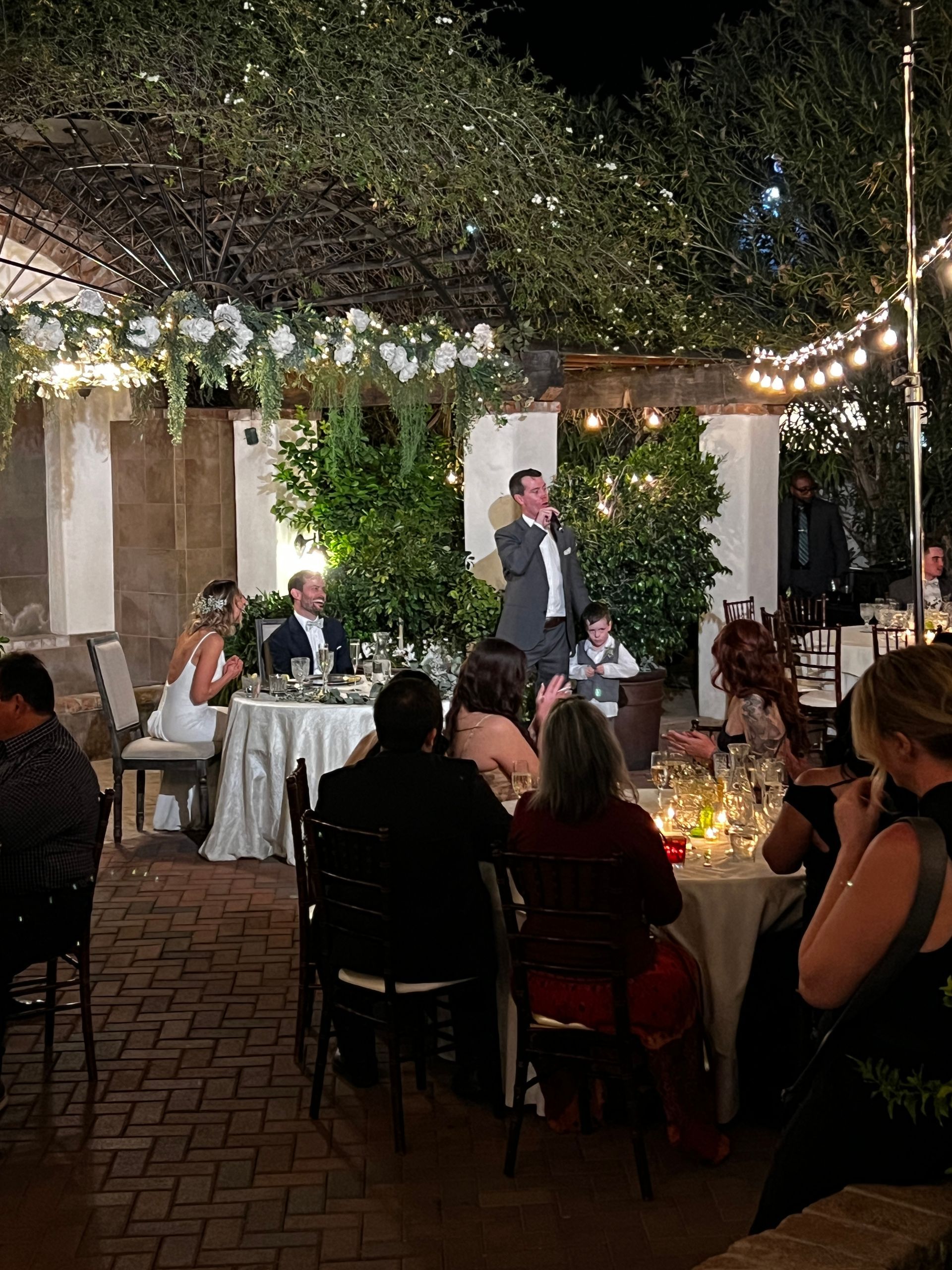 A man giving a speech at an outdoor wedding reception. Guests sit at tables, and string lights and greenery decorate the space.