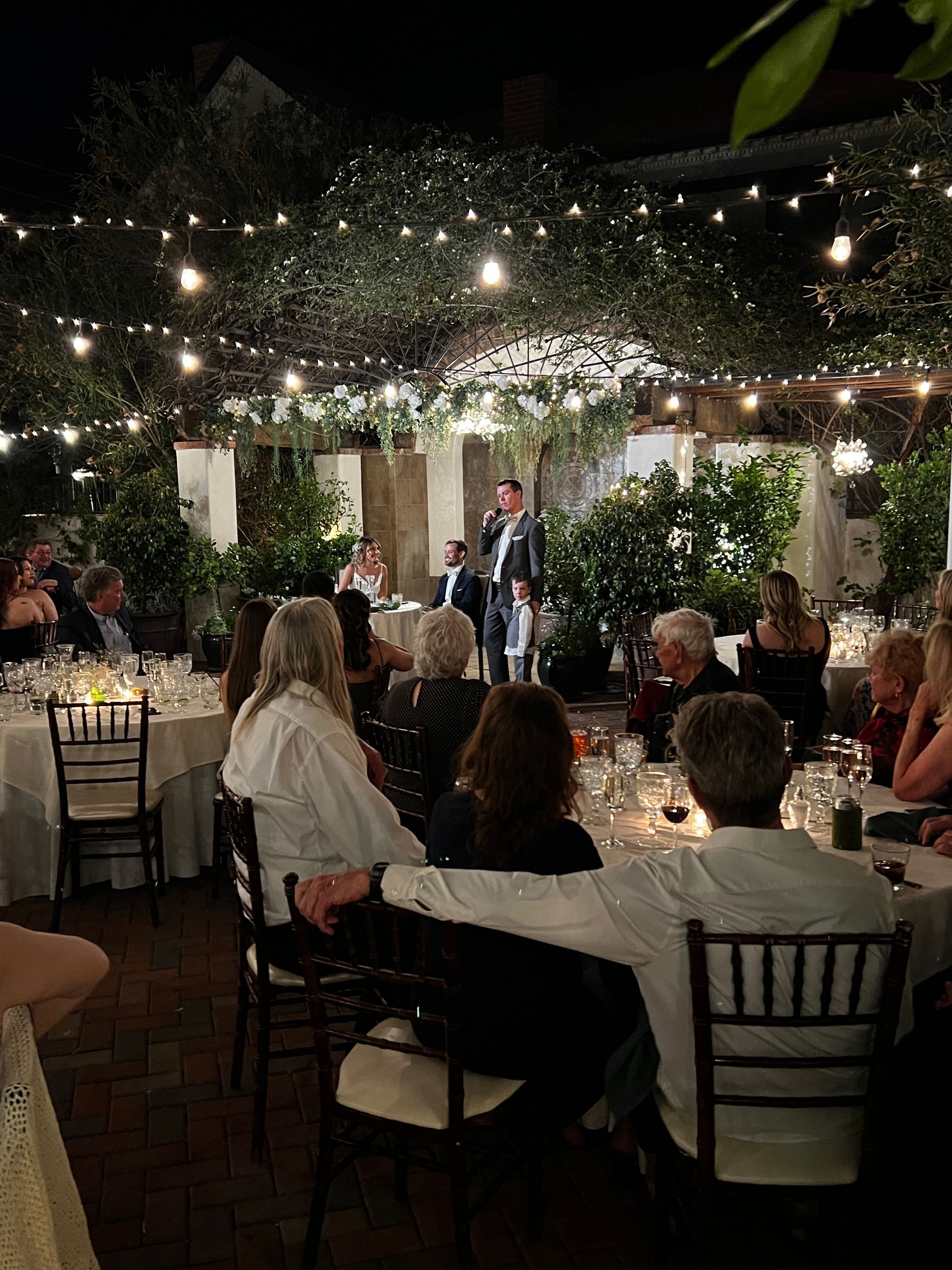 People seated at tables listen to a man speaking at an outdoor event under string lights.