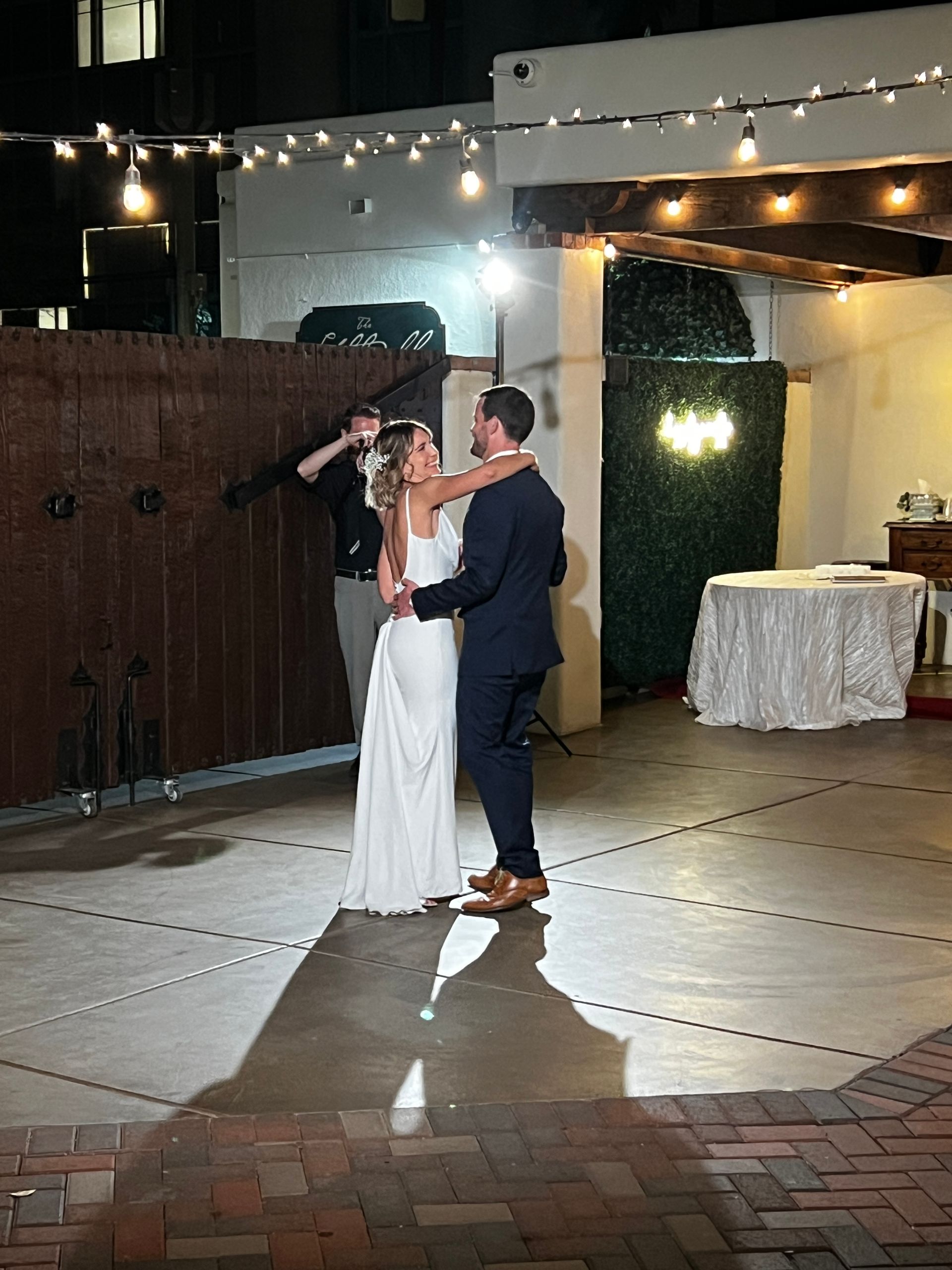 Bride and groom dancing outdoors at night, under string lights. A photographer captures them with a flash.