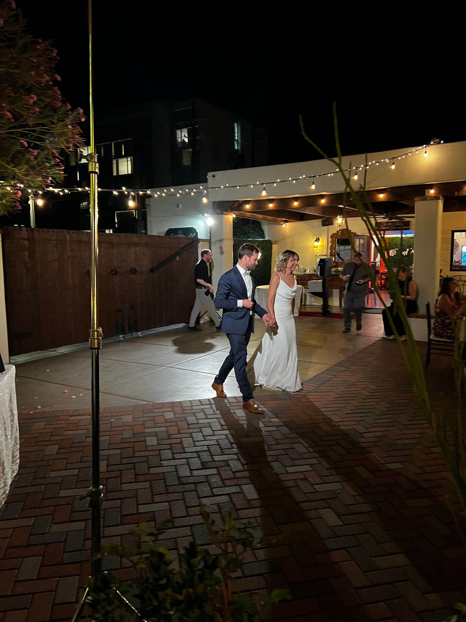 Bride and groom walk hand-in-hand at night, exiting a wedding venue. Brick patio with string lights and guests in background.