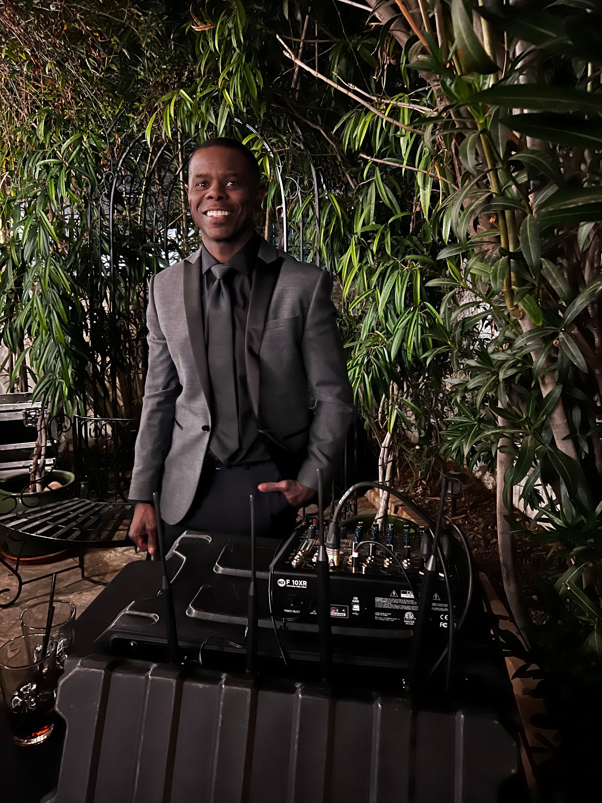 Man in a gray suit smiling, standing behind a Theremin instrument in an outdoor setting with lush greenery.