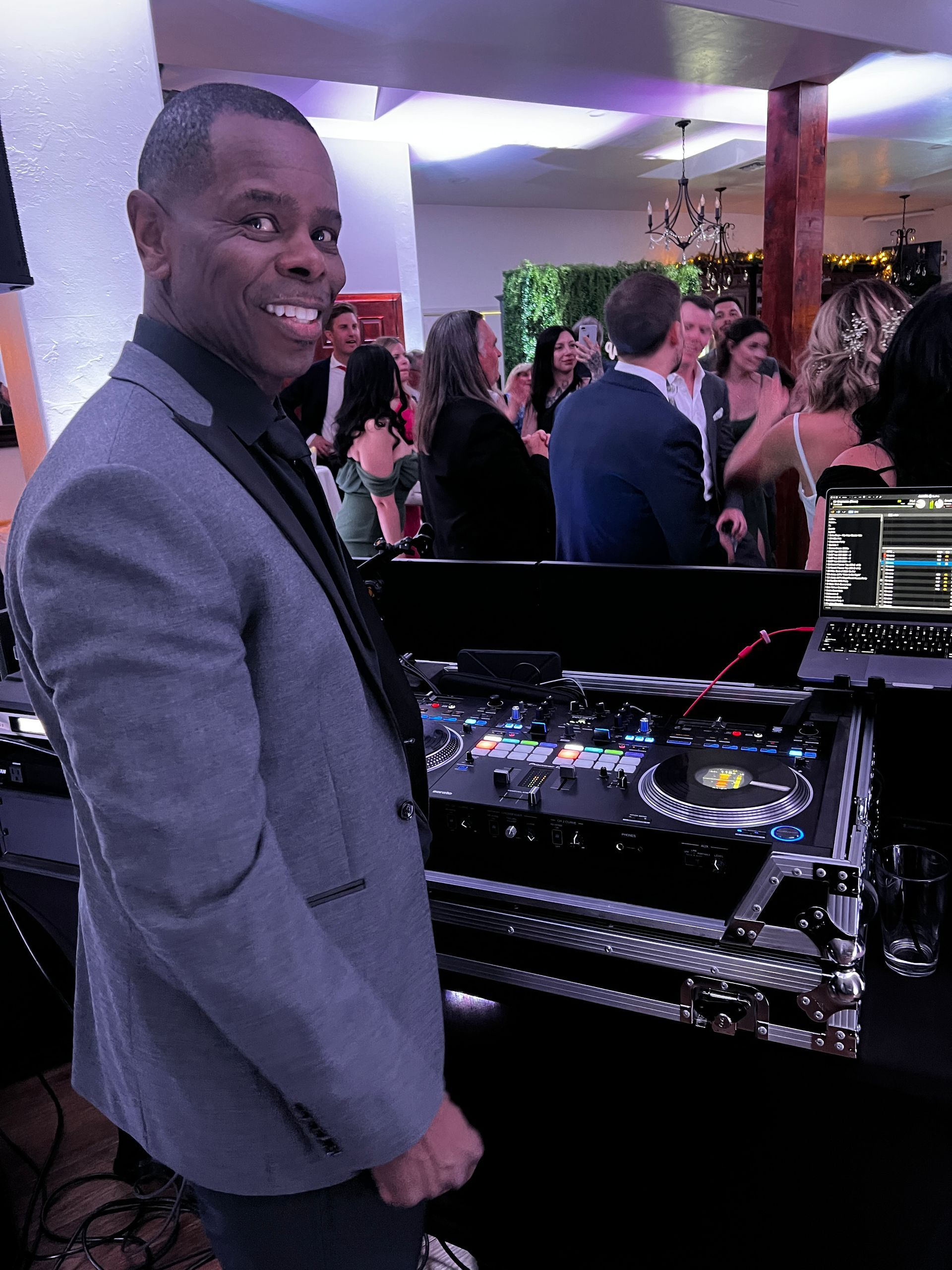 Man smiling, DJing at an event. He wears a gray blazer. A crowd and equipment are visible in the background.