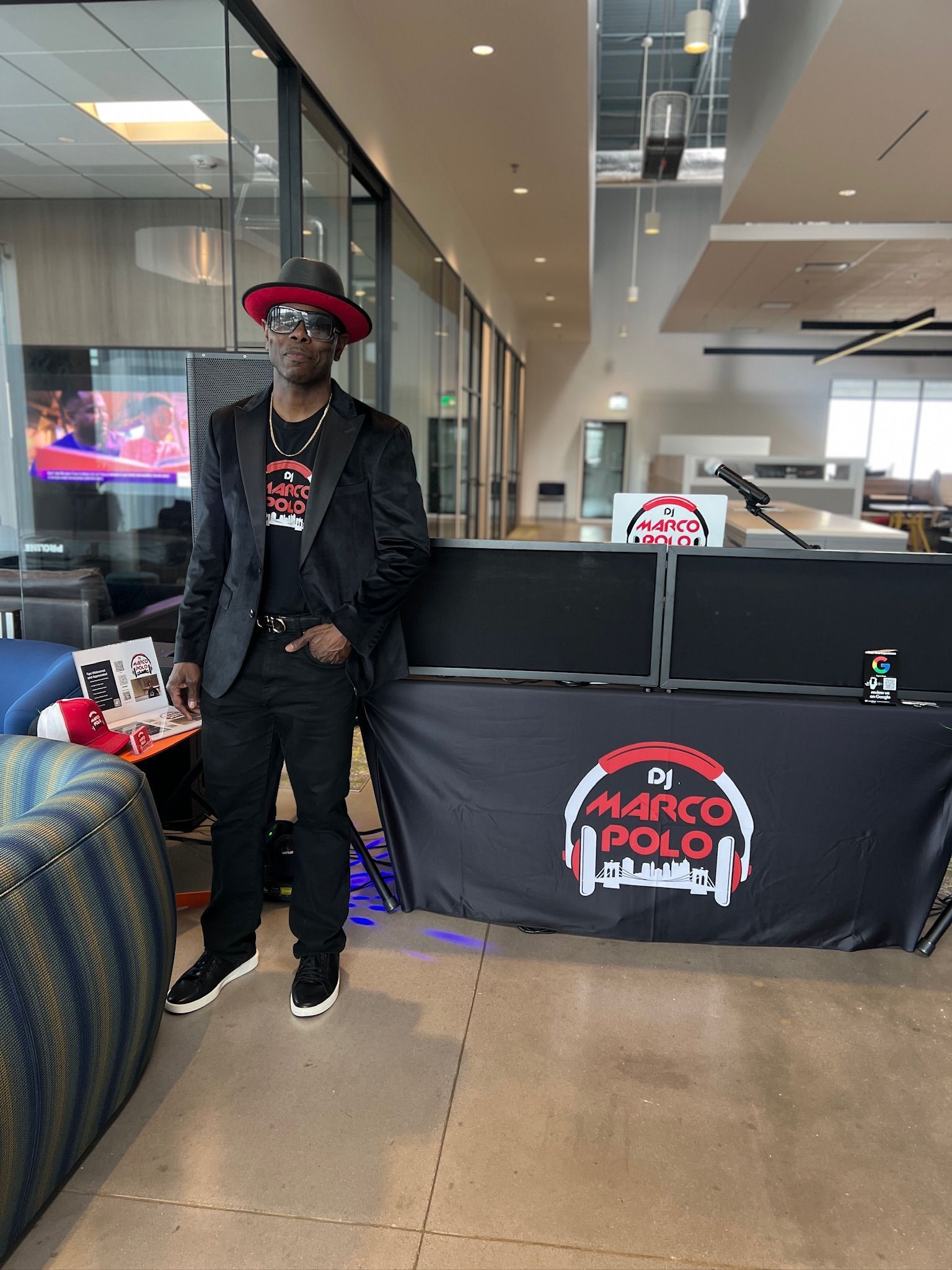 DJ Marco Polo stands at his DJ setup indoors, wearing a black blazer and a red-brimmed hat. His logo is visible on the table.