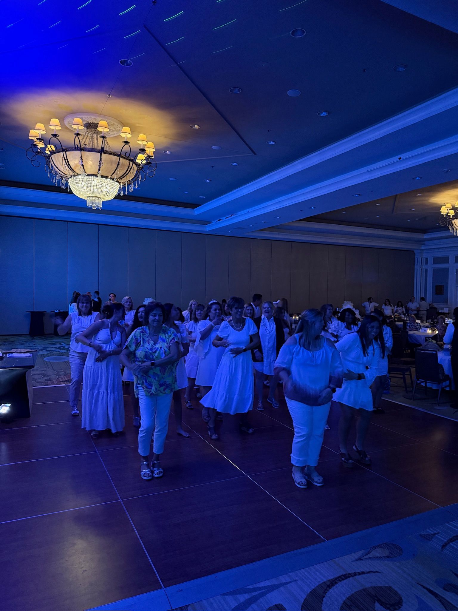 People dancing in a ballroom lit with blue light, with a chandelier overhead. Most wear white outfits.