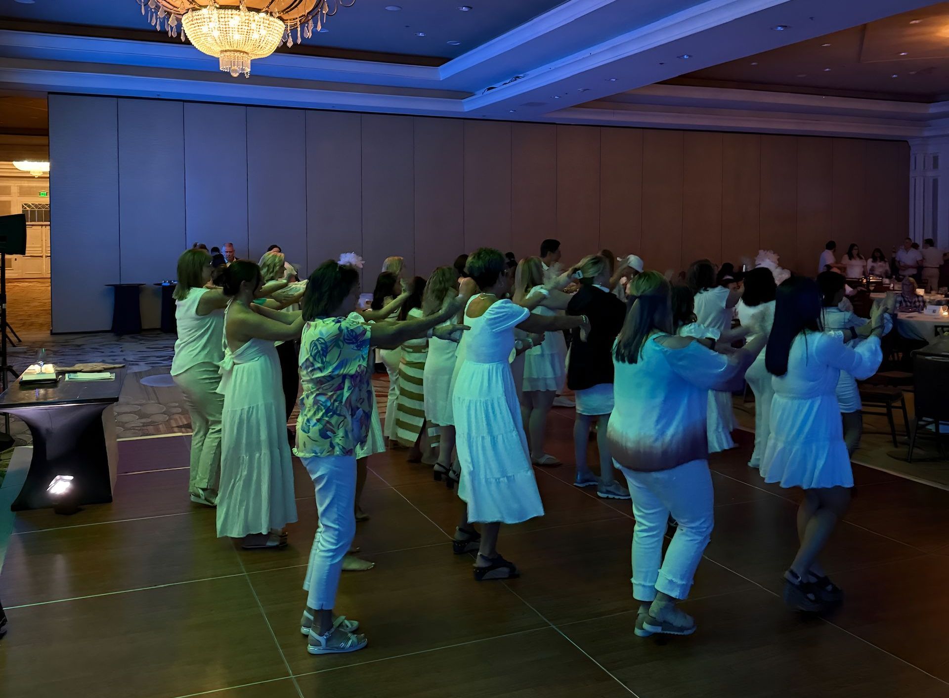 A group of people in a ballroom dancing, most in white clothing. Overhead lighting creates a blue and gold ambiance.