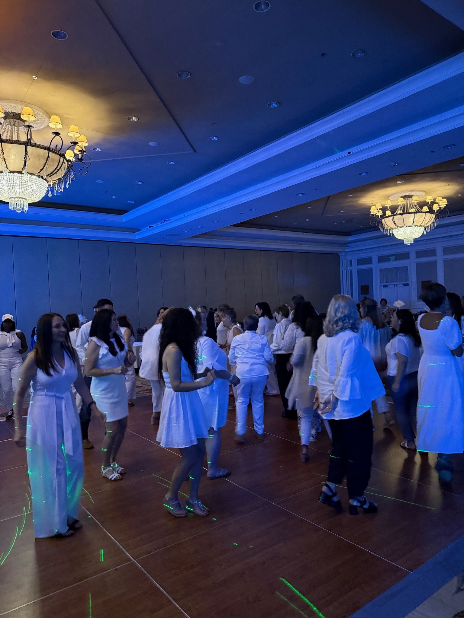 People in white outfits dance in a large room with blue lighting. Chandeliers hang from the ceiling.
