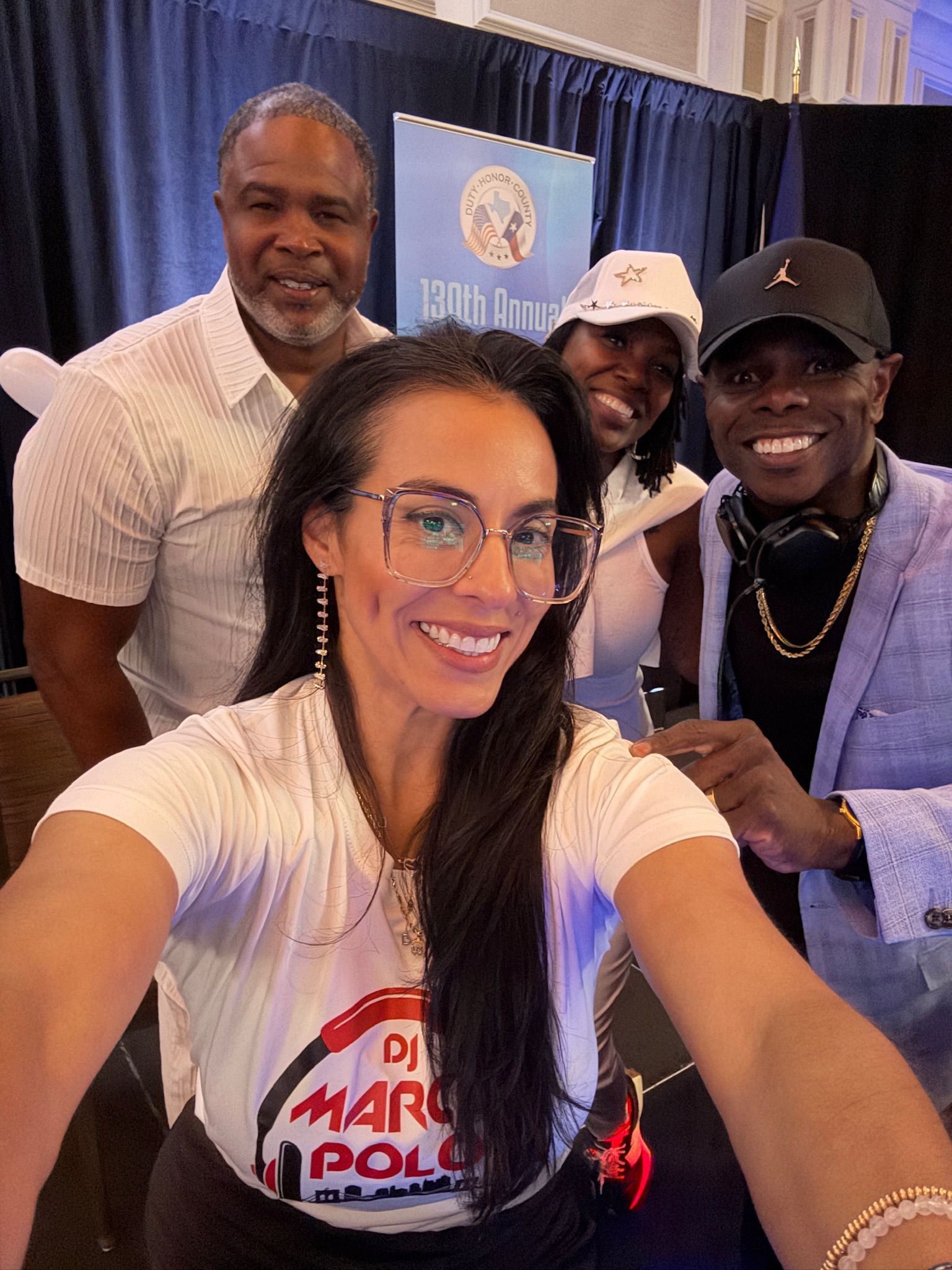 Group selfie: Four people smiling. Woman in glasses takes photo, DJ in blazer, others in background.