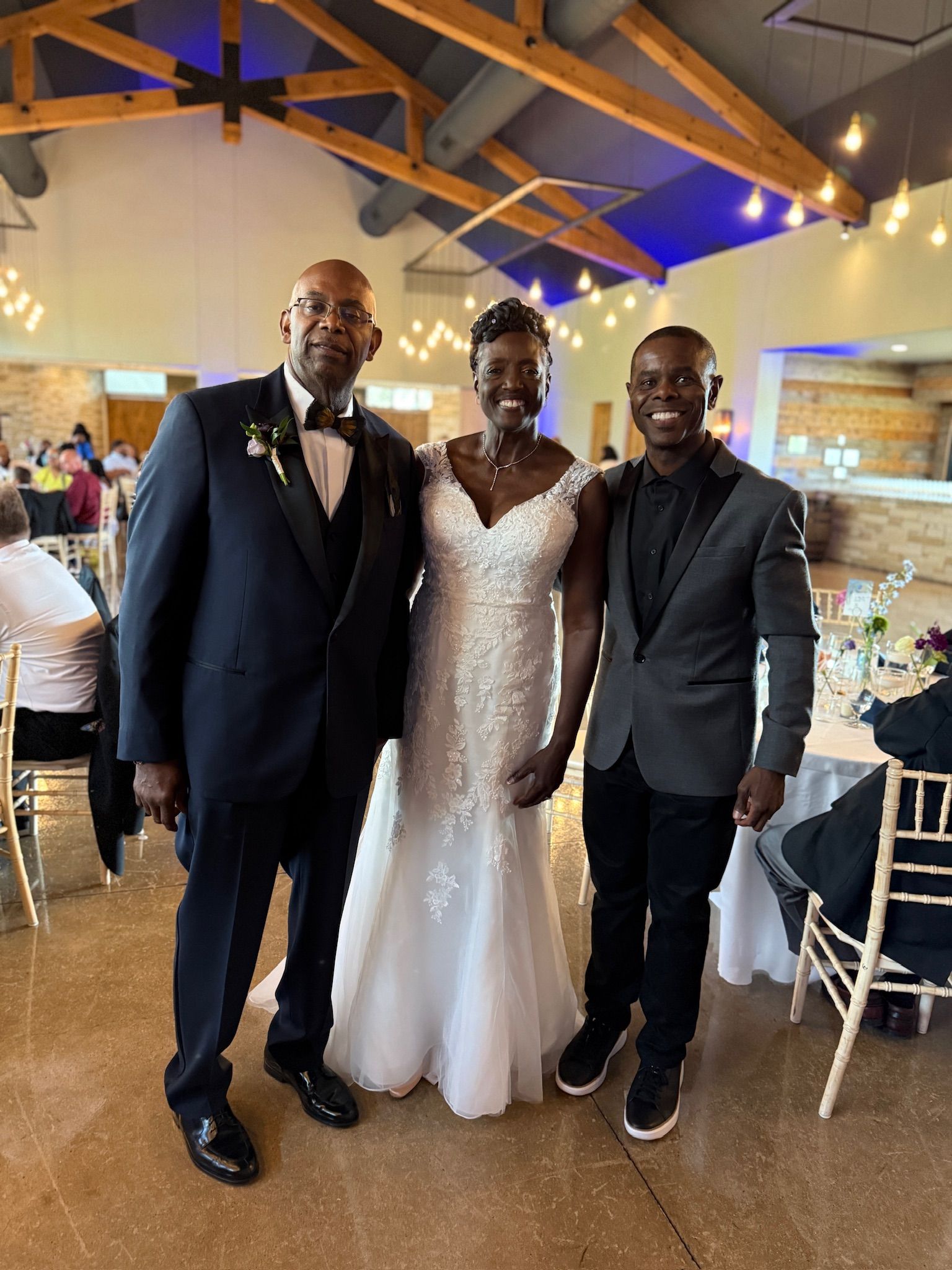 A bride in a white wedding gown stands between a man in a tuxedo and another in a black blazer. They are indoors at a reception.